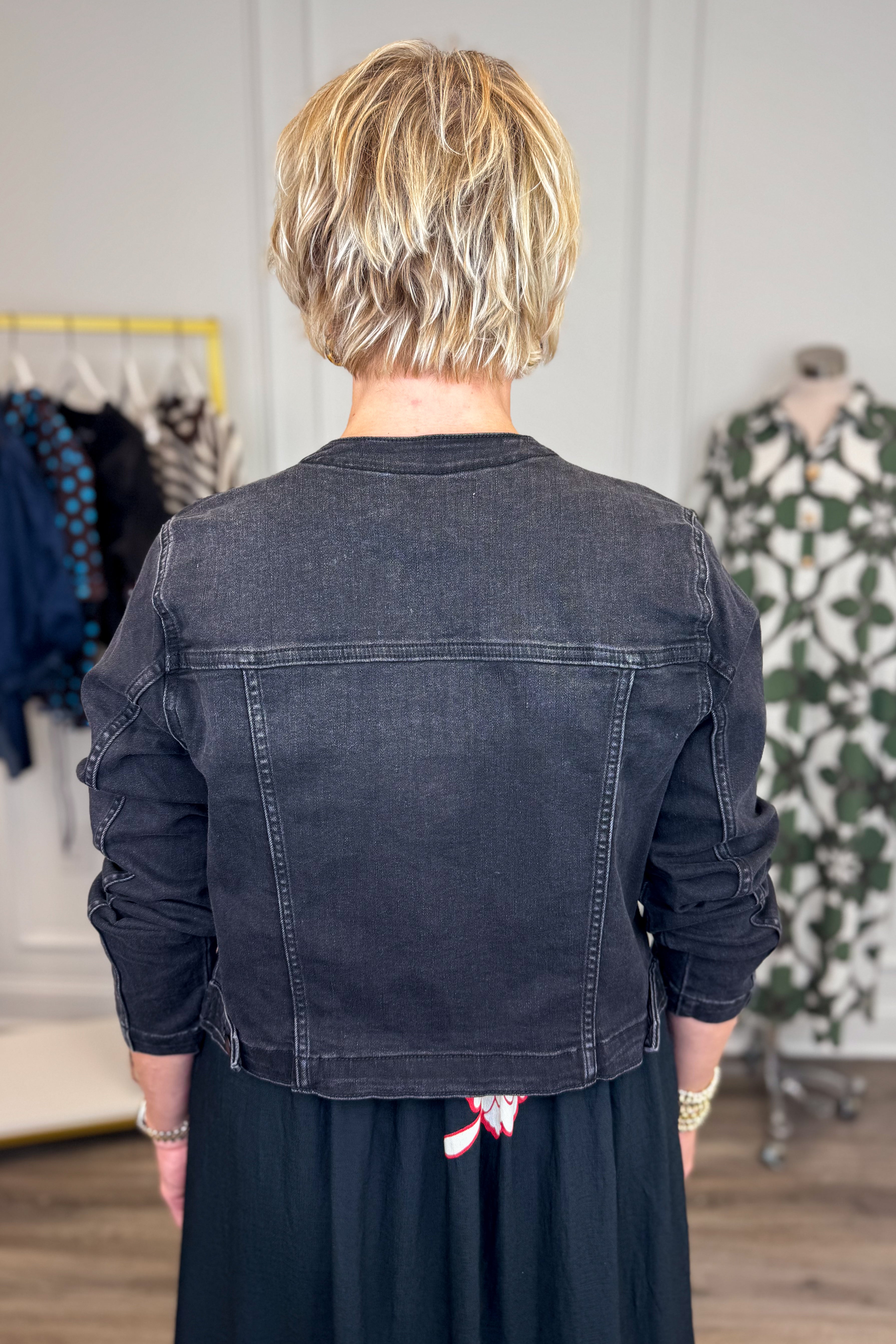 Back view of woman wearing Swagger Boutiqueās washed black denim jacked with dress with clothing hanging in the background.