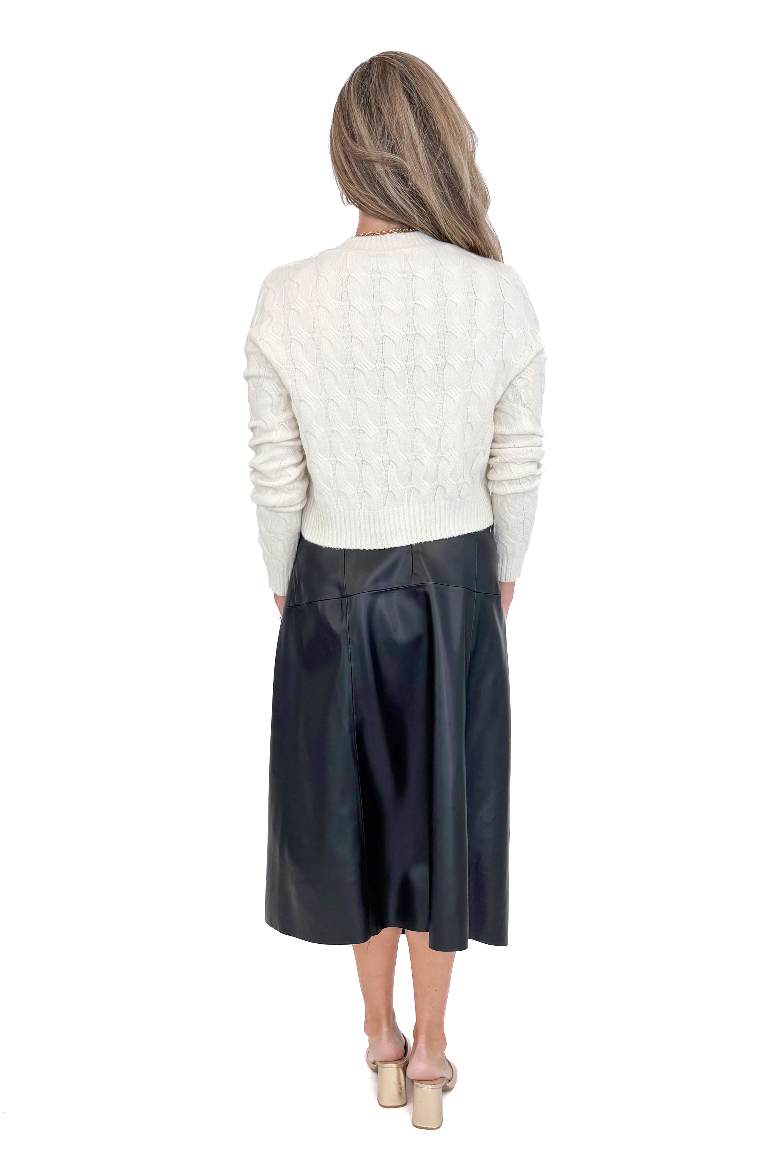 Back view of woman wearing cream sweater with black faux leather skirt against white studio background.