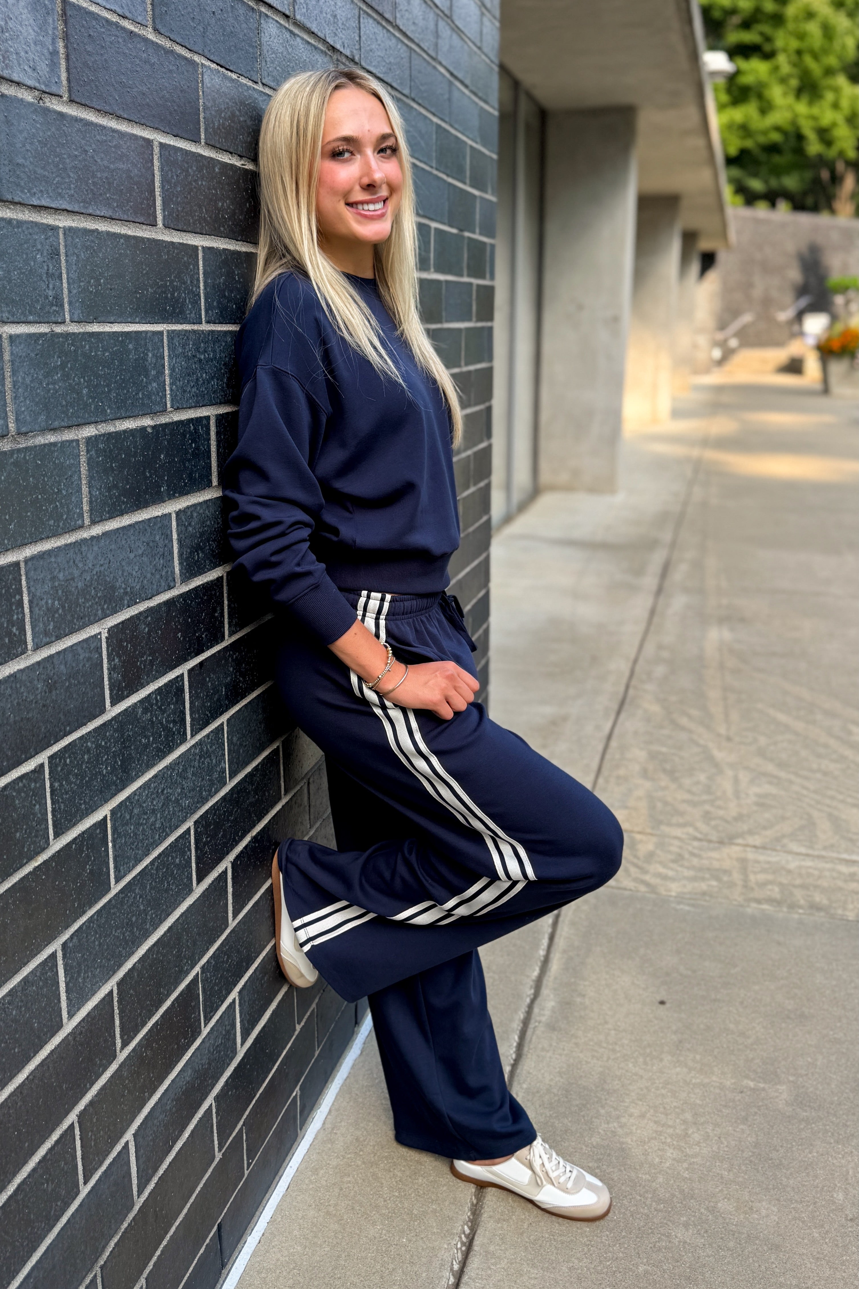 Front view of woman wearing Swagger Boutiqueās Eclipse Breakaway Track Pants by Z Supply with Eclipse Comeback Sweatshirt by Z Supply leaning against a brick wall outside.