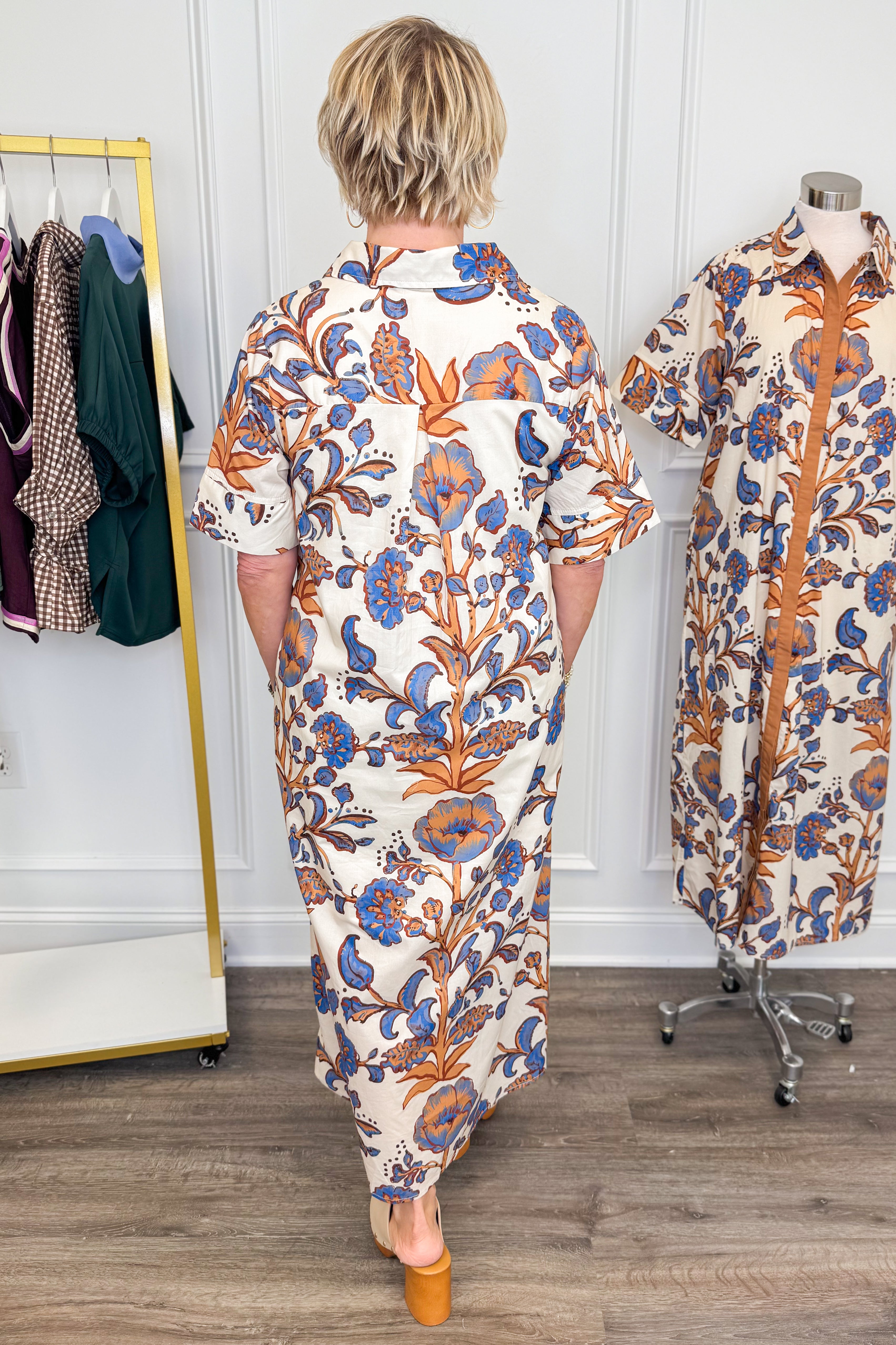 A person viewed from behind wearing a cream shirt dress with a blue and orange floral print, standing in a boutique next to a mannequin displaying the same dress.