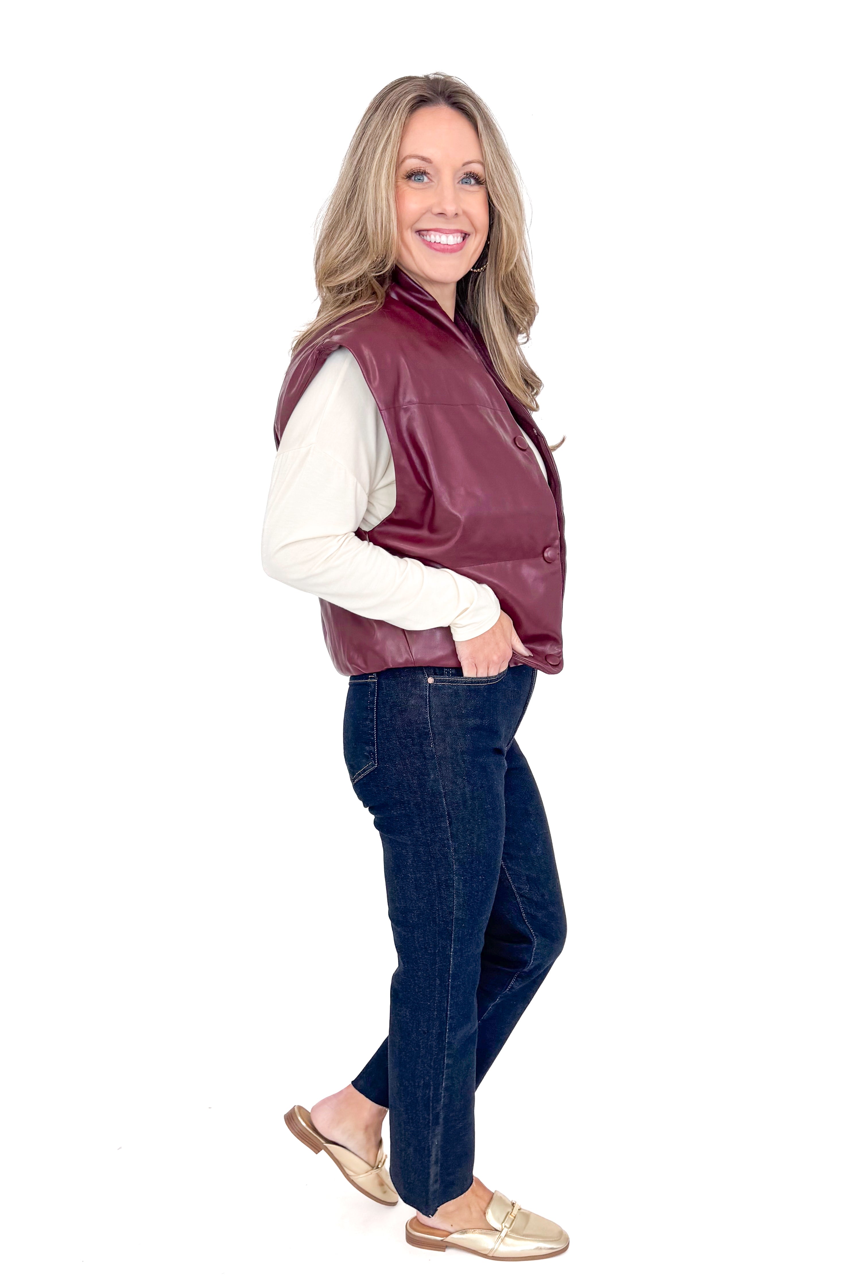 Side view of woman wearing plum faux leather vest with jeans against white studio background.