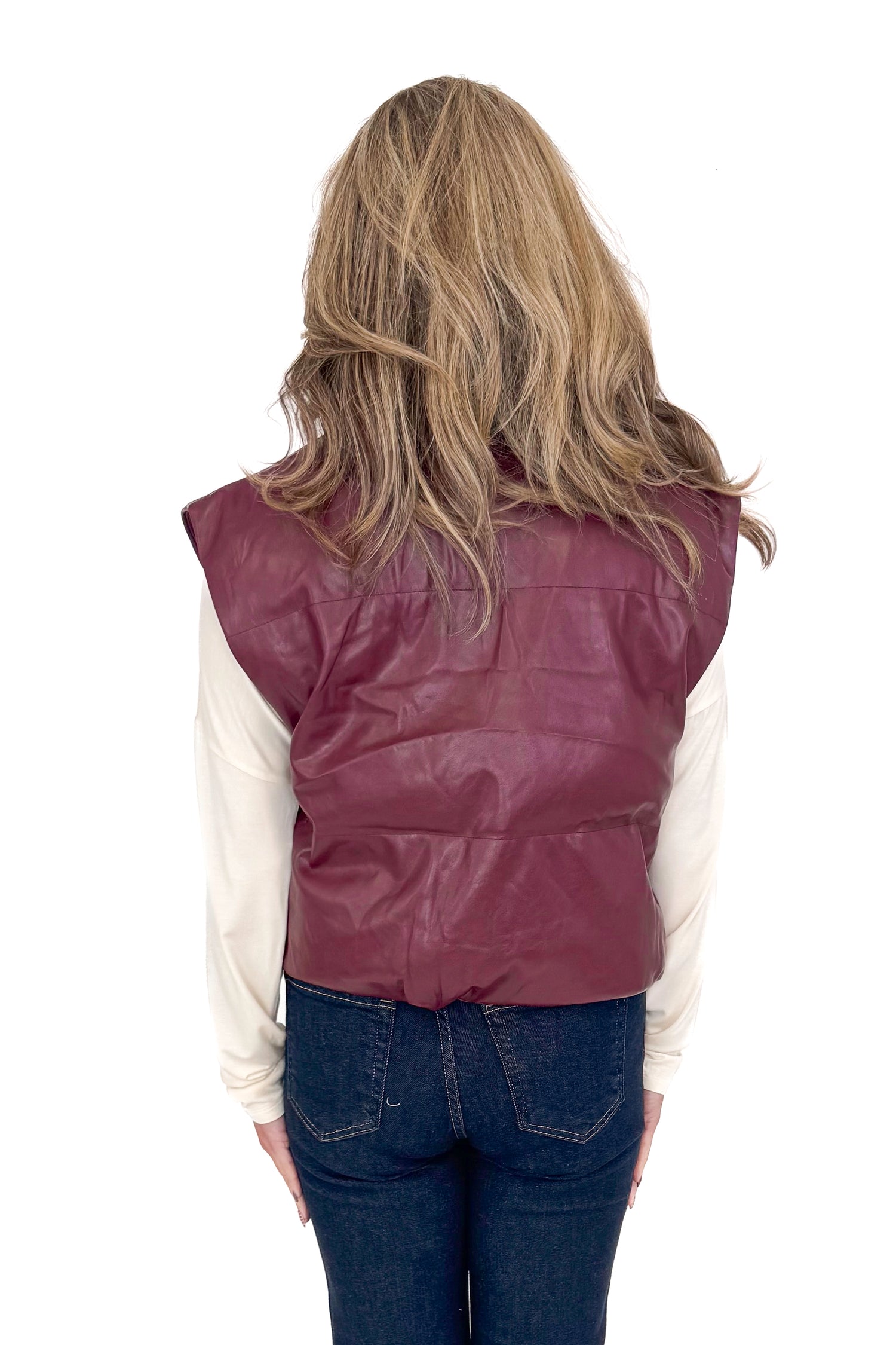 Back view of woman wearing plum faux leather vest with jeans against white studio background.