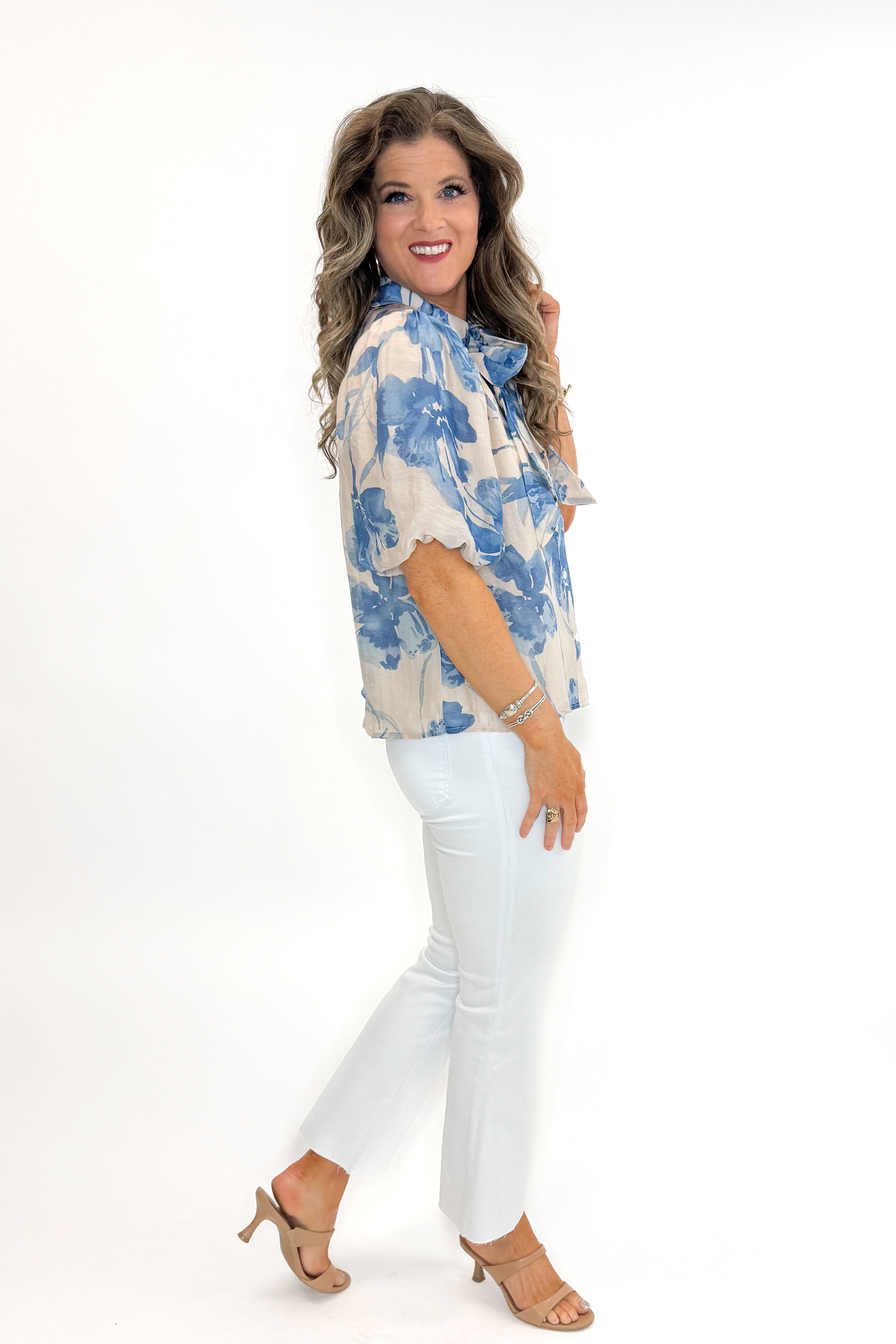 Side view of woman wearing Cream & Blue Floral Neck Tie Detail Top by Fate with white jeans against white studio background.