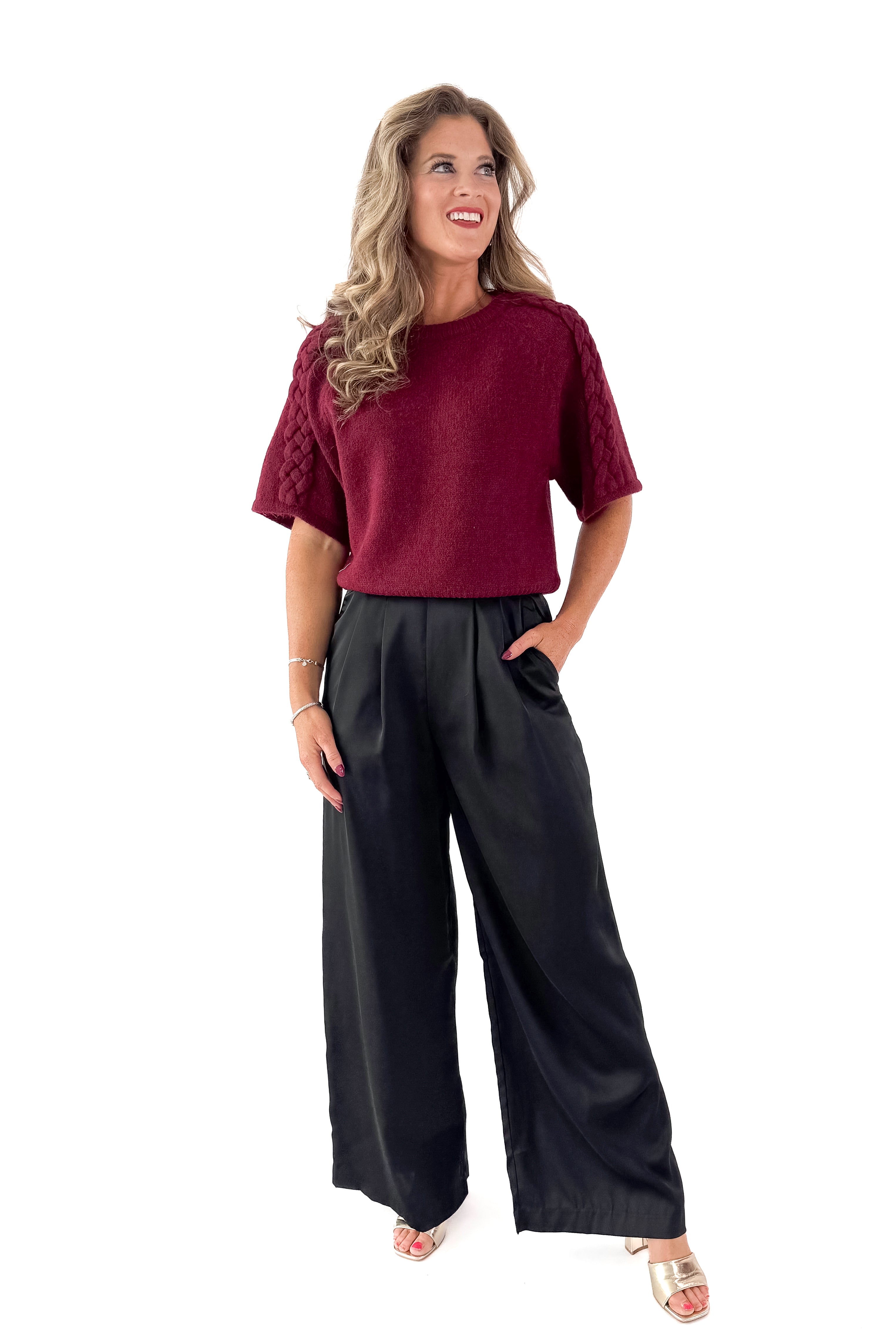 Front view of woman wearing sweater with black satin pleated pants against white studio background.