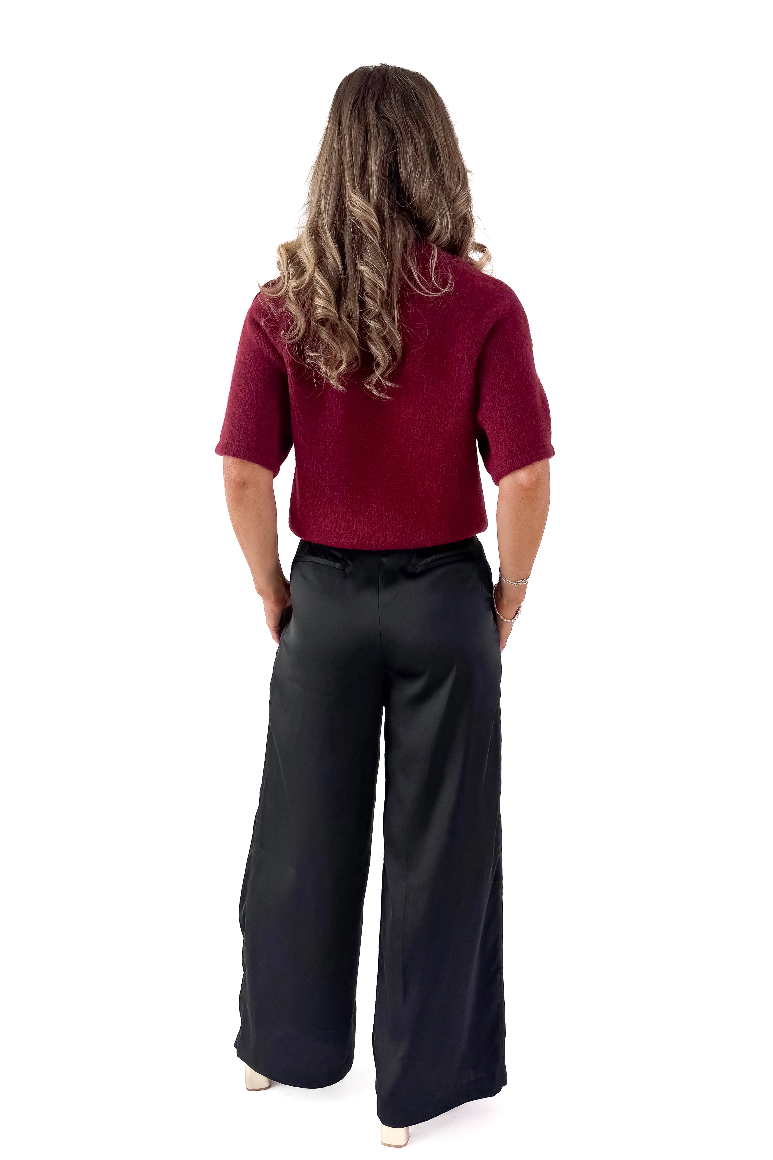 Back view of woman wearing sweater with black satin pleated pants against white studio background.