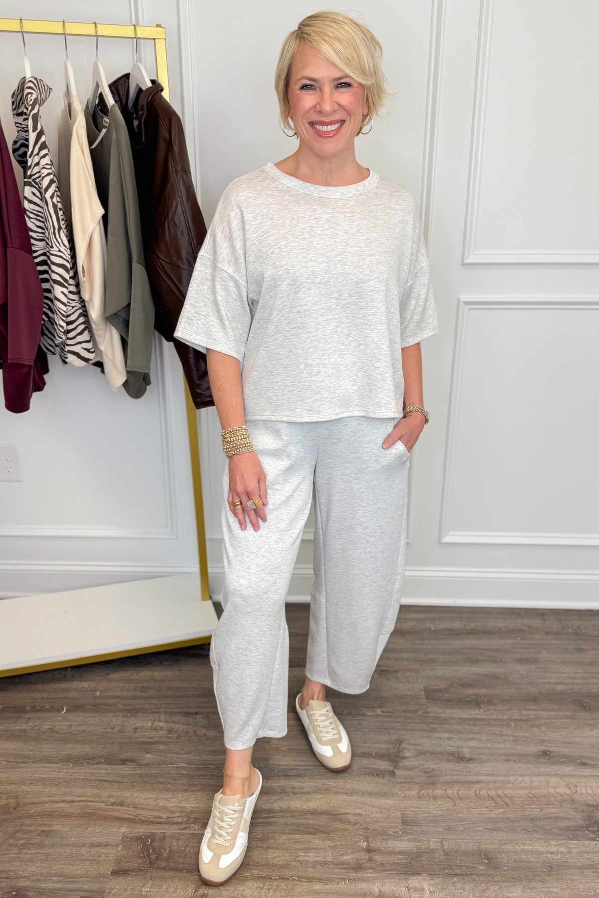 Front view of woman wearing Swagger Boutique’s Grey Round Neck Short Sleeve Top with matching barrel pants with clothes hanging in the background.