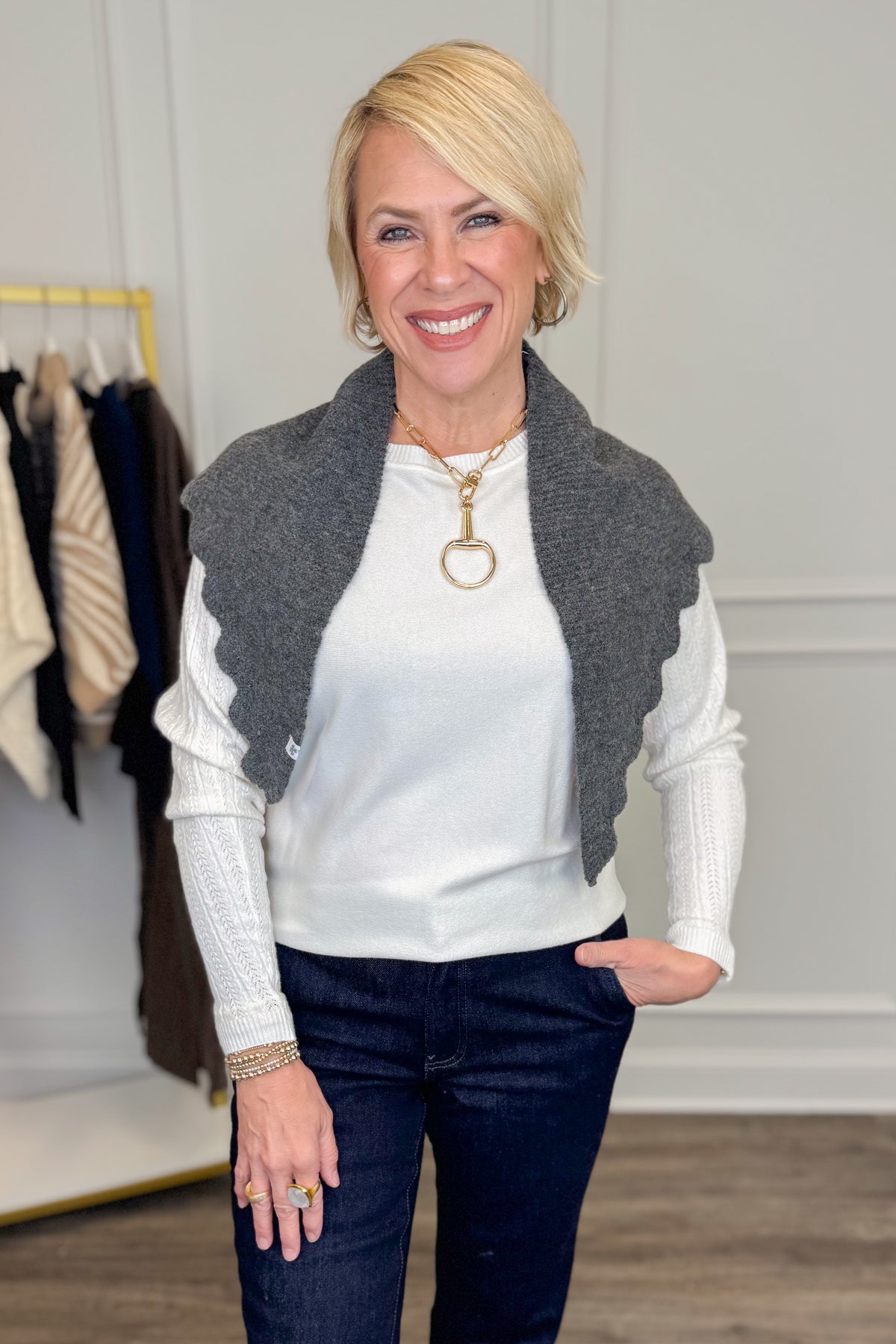 A smiling blonde woman wearing a white ribbed long-sleeve top, dark blue jeans, and a gray scalloped knit scarf, standing in a boutique setting with a clothing rack visible in the background.