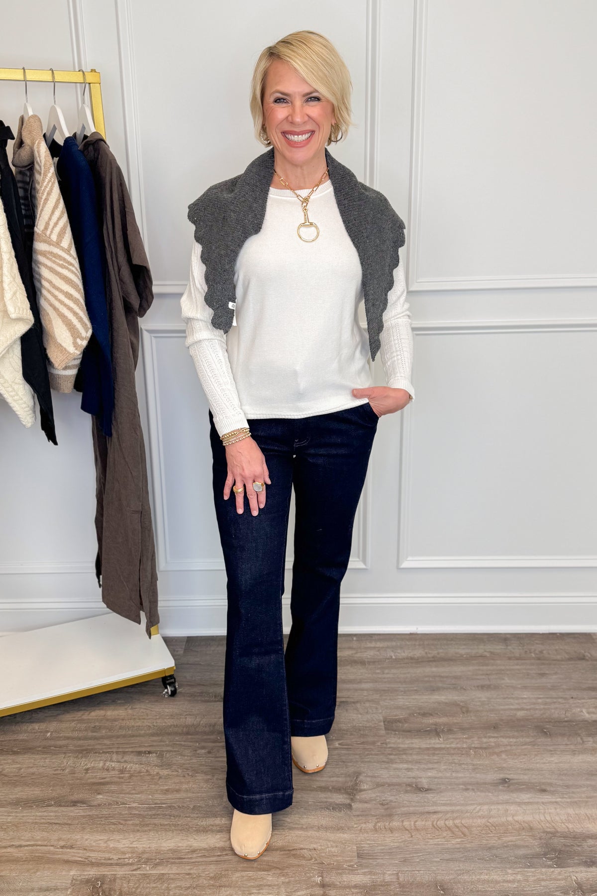 A woman modeling a white knit top with a dark gray ruffled scarf paired with dark blue flare jeans and beige heels, standing in a room with a clothing rack and paneled wall.