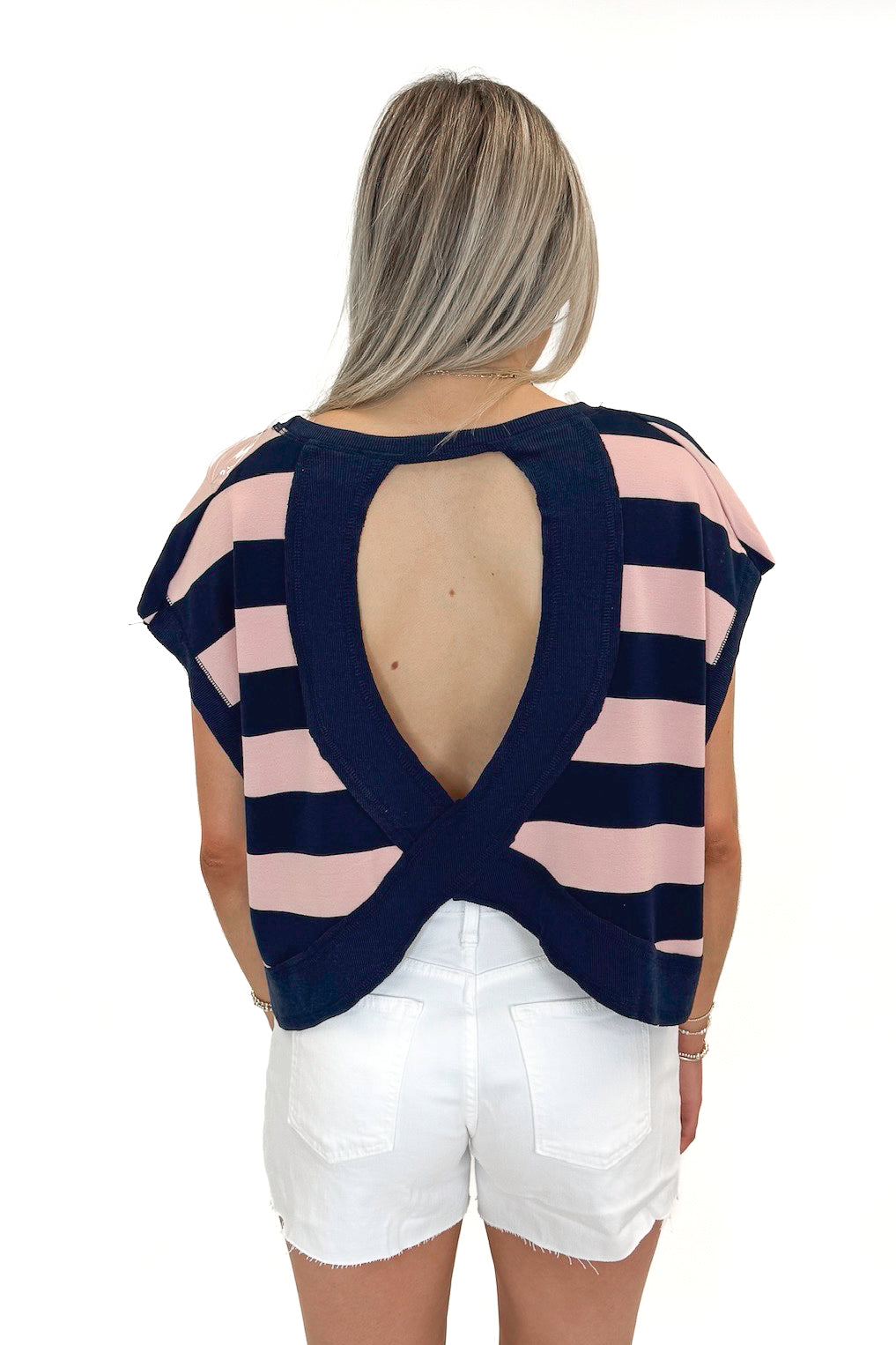 Back view of woman wearing Navy & Pink Striped Cut Out Back Tee with jean shorts against white studio background.
