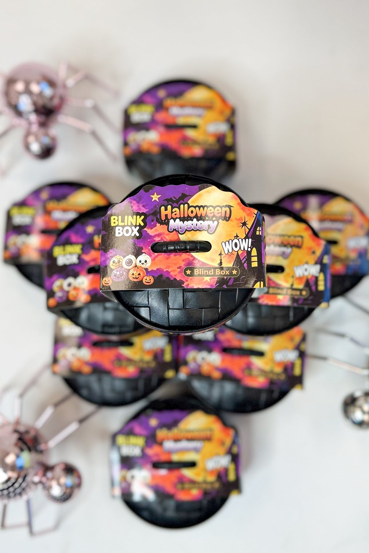 Several Halloween-themed blind box packages arranged in a circle on a white surface, with spider decorations in the corners. The front package is most prominent and shows the text &