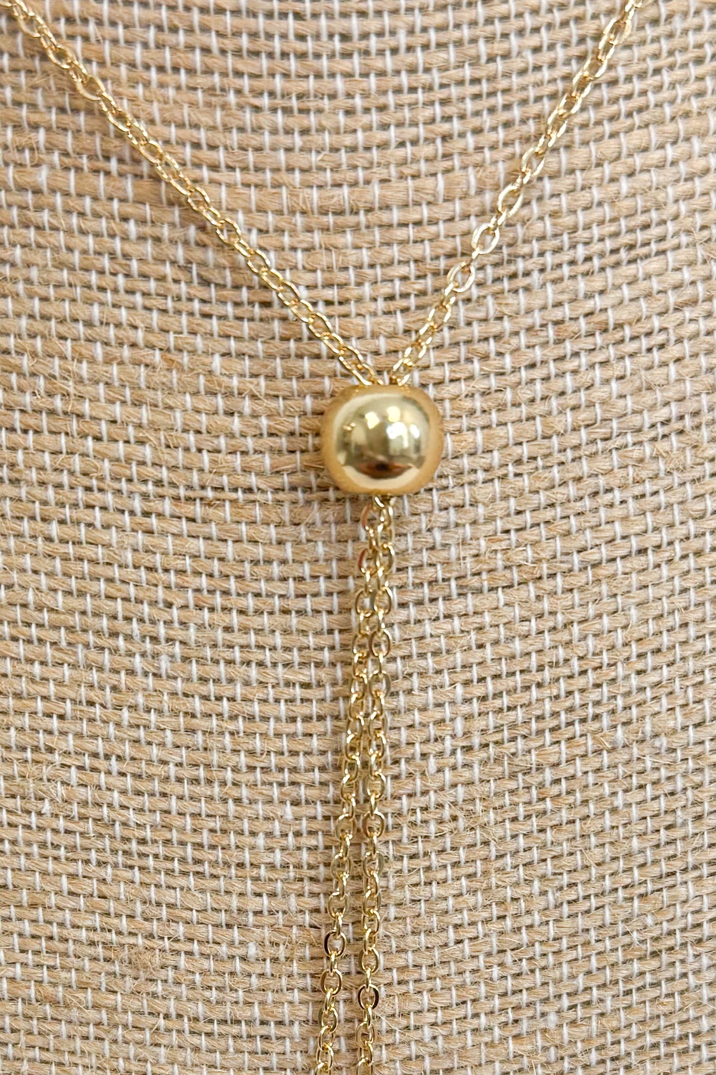 Close up of gold-tone bolo necklace featuring a fine, reflective chain held together by a smooth, polished gold sphere slider