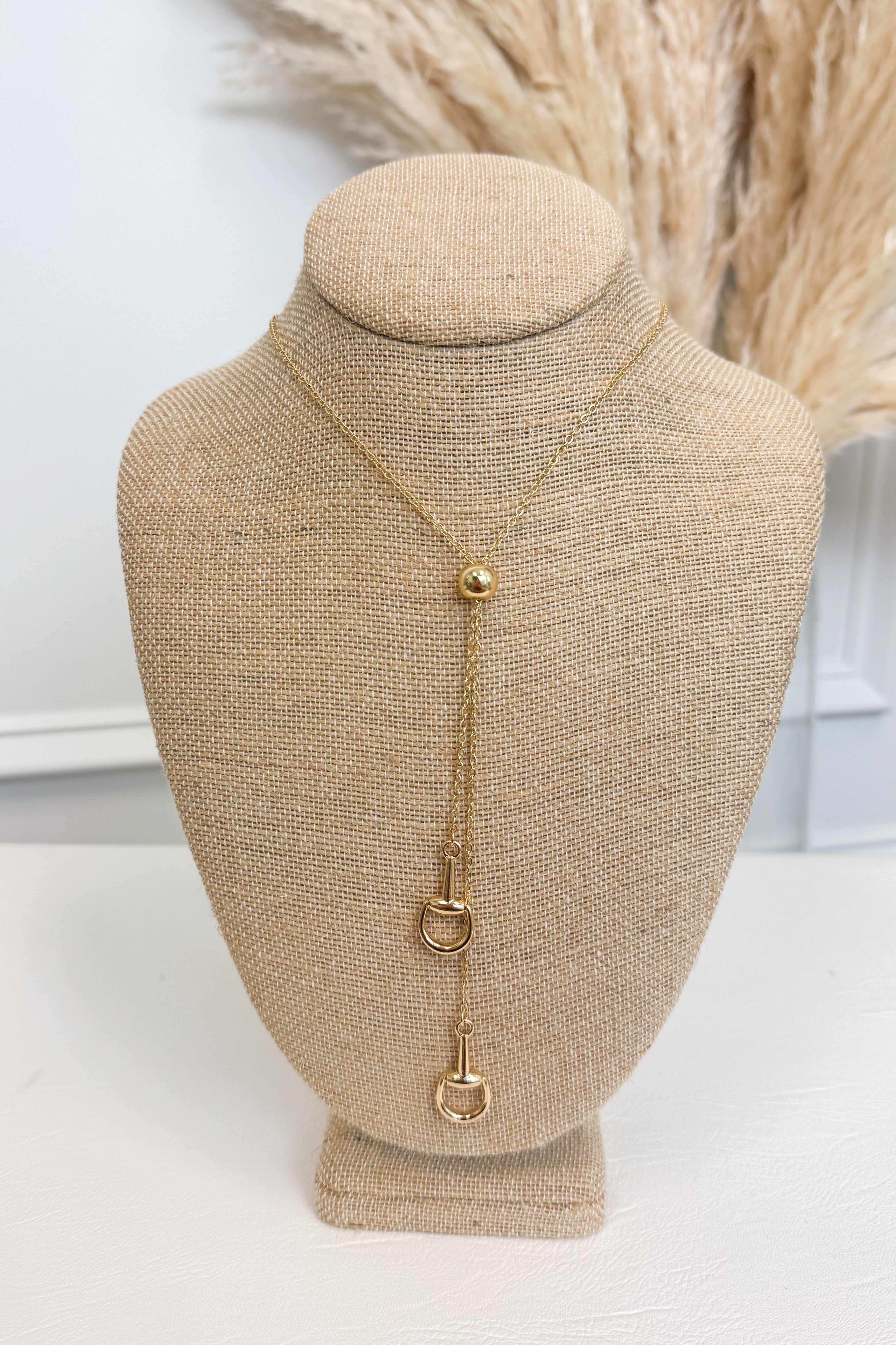 Gold-tone bolo necklace featuring a fine, reflective chain held together by a smooth, polished gold sphere slider, with delicate chain strands dropping vertically down the center each with a small horseshoe pendant