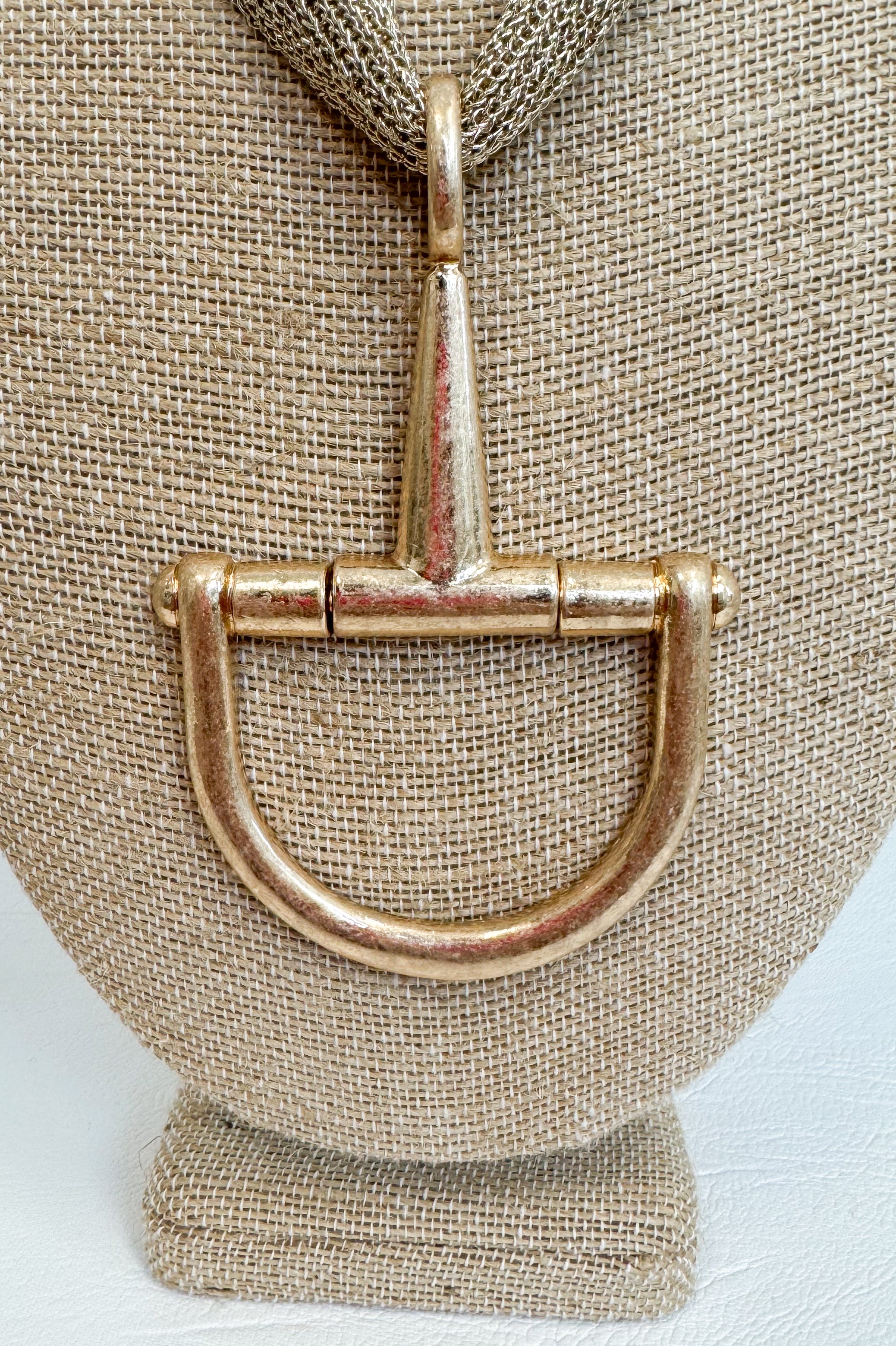 Close up of gold-tone mesh necklace featuring a large horsebit pendant, displayed on a beige jewelry bust against a white studio background