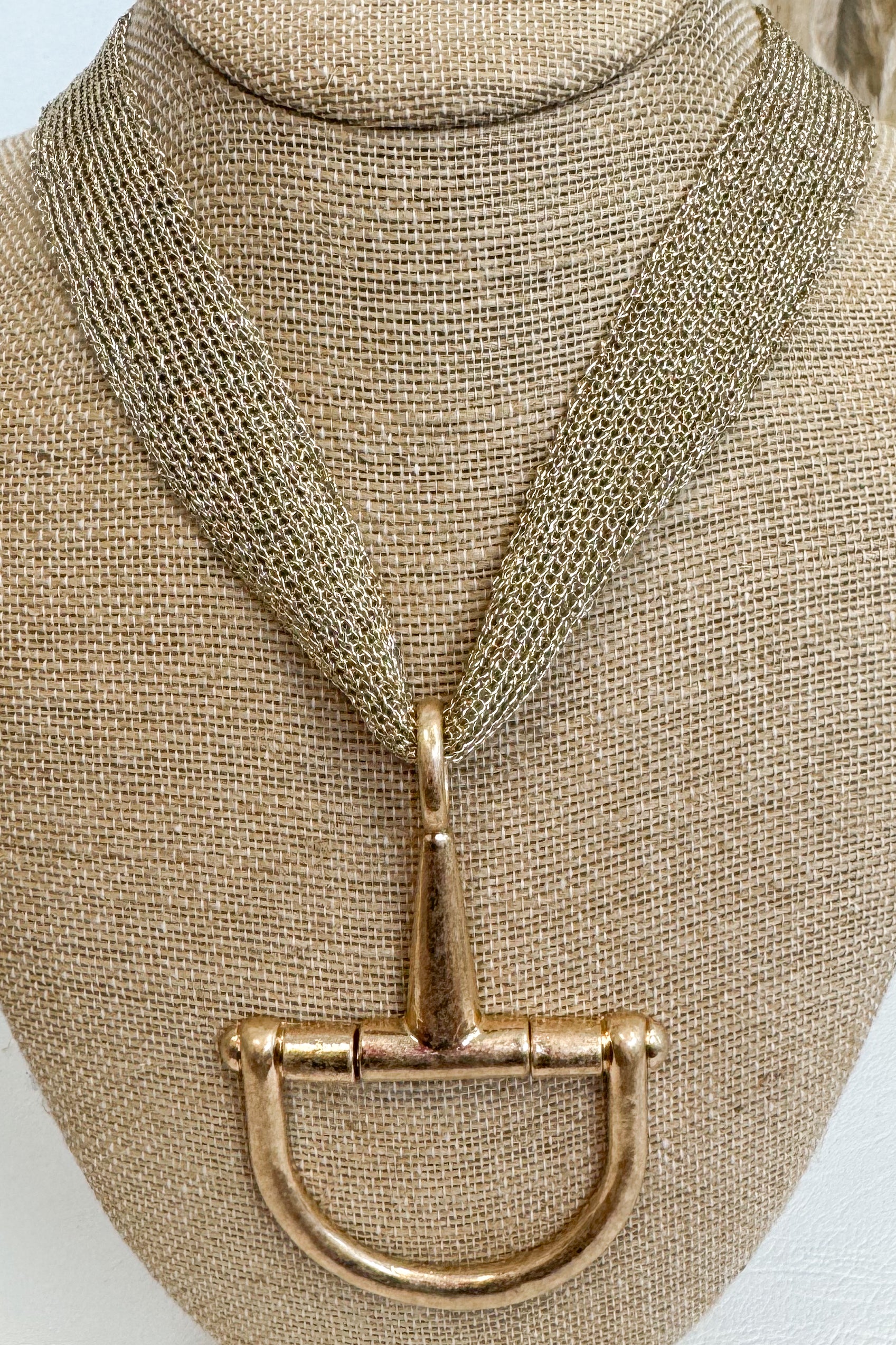 Gold-tone mesh necklace featuring a large horsebit pendant, displayed on a beige jewelry bust against a white studio background