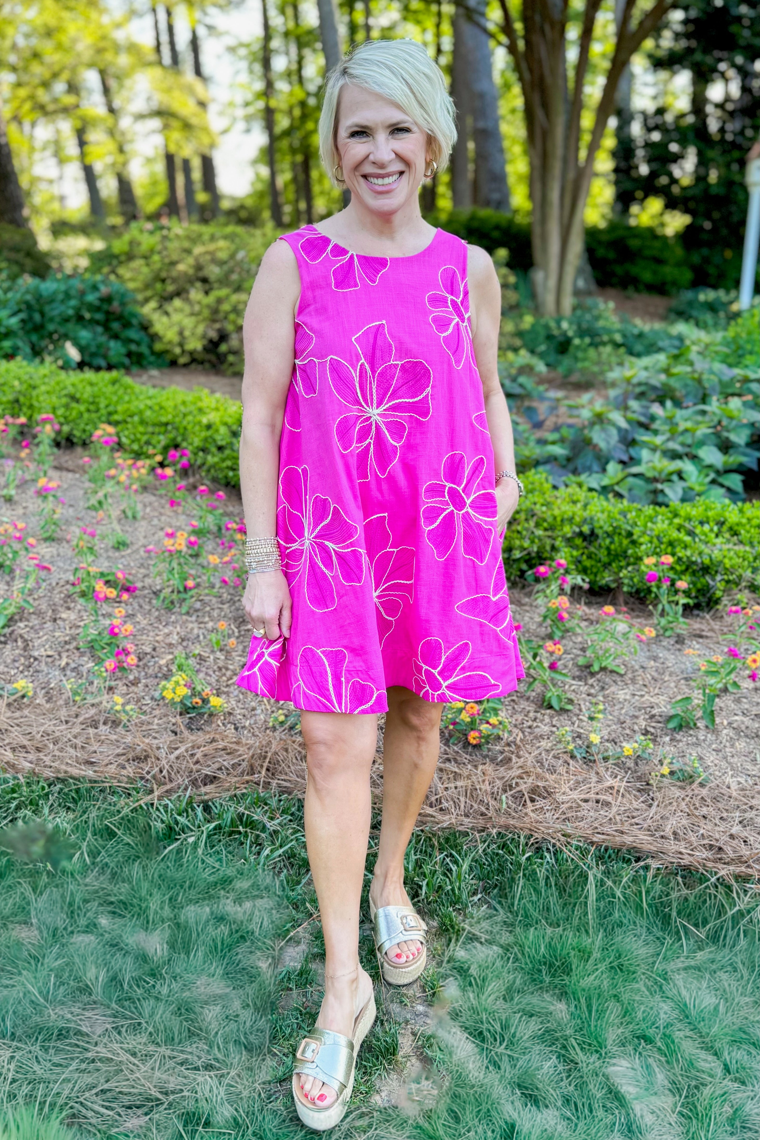 Front view of woman wearing Swagger Boutique’s Hot Pink Floral Embroidered Mini Dress with sandals outside at a park with flowers.