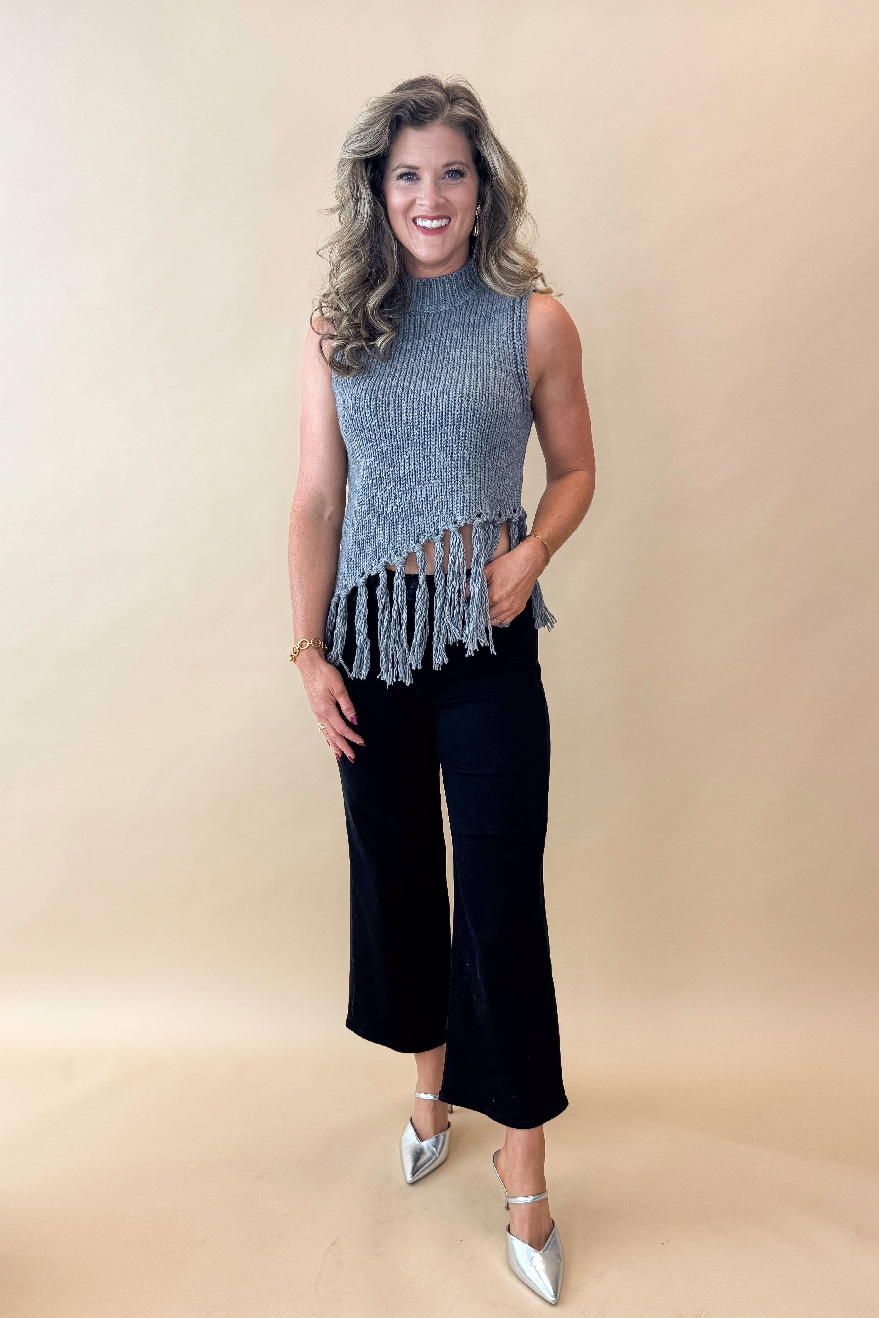 Front view of woman wearing Grey Mock Neck Fringe Detail Sleeveless Sweater and jeans against tan studio background.