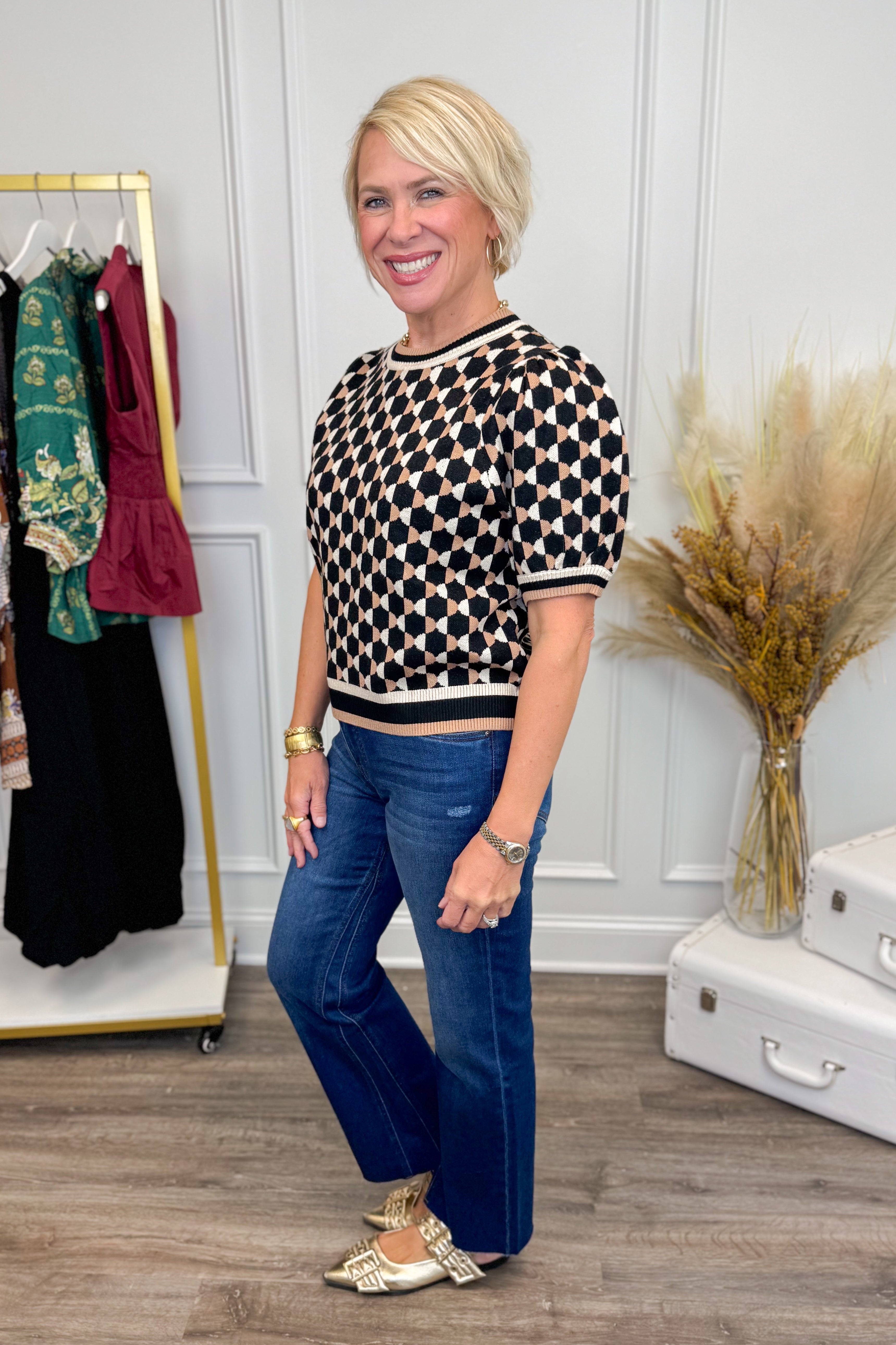 Woman wearing a patterned top and blue jeans standing in a room with a rack of clothes and decorative elements.