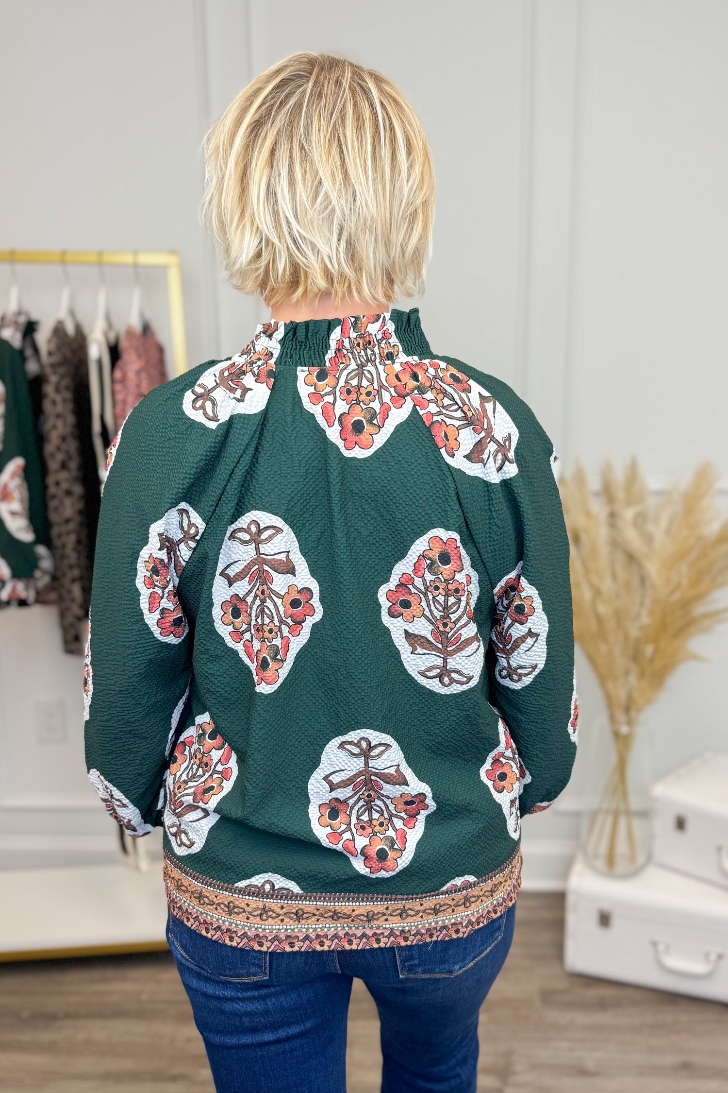 Back view of Woman wearing Hunter Green Printed & Textured Puff Sleeve Top and jeans with clothes in background.