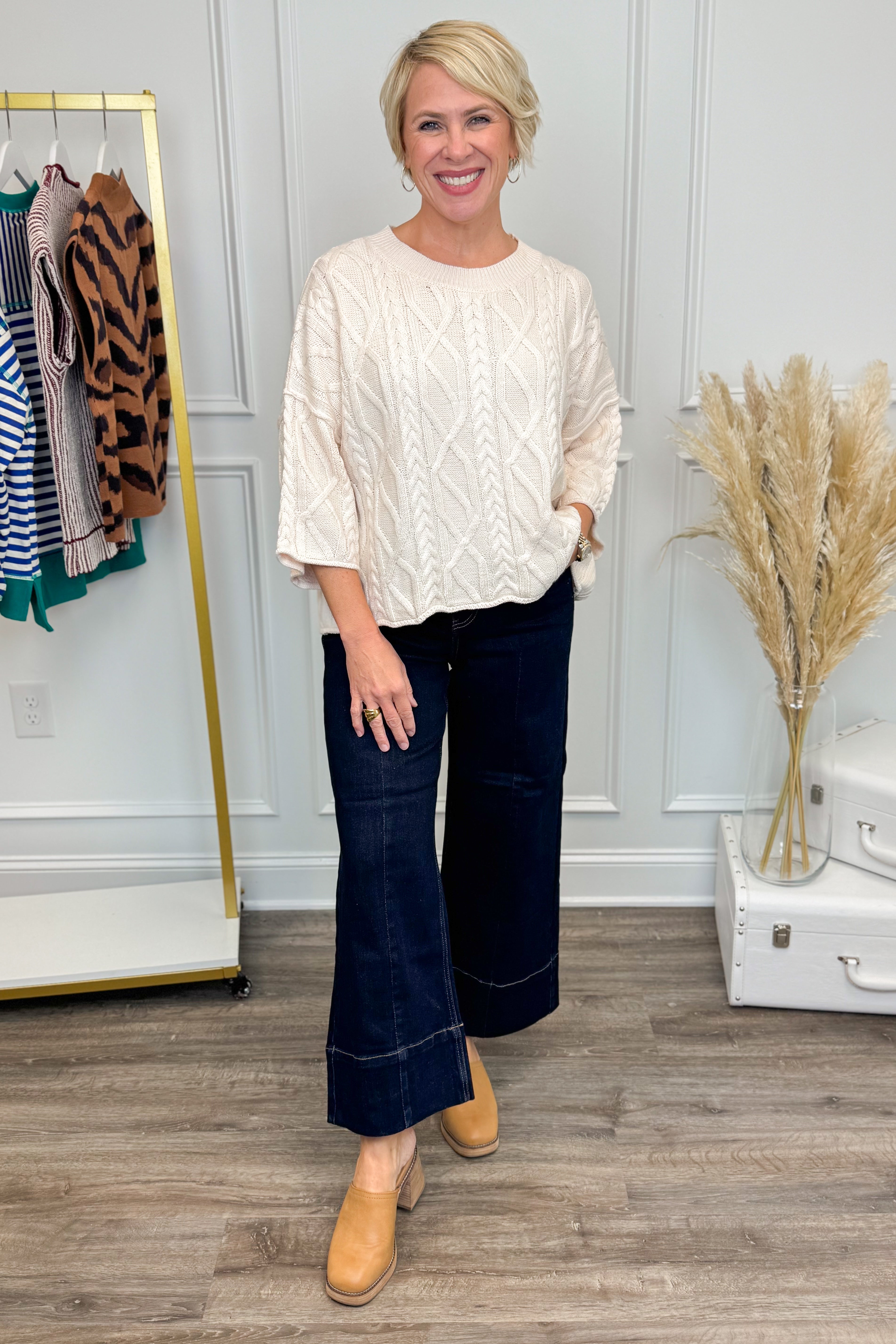 Woman in a cream blouse and dark jeans standing in a room with a clothing rack and decor.
