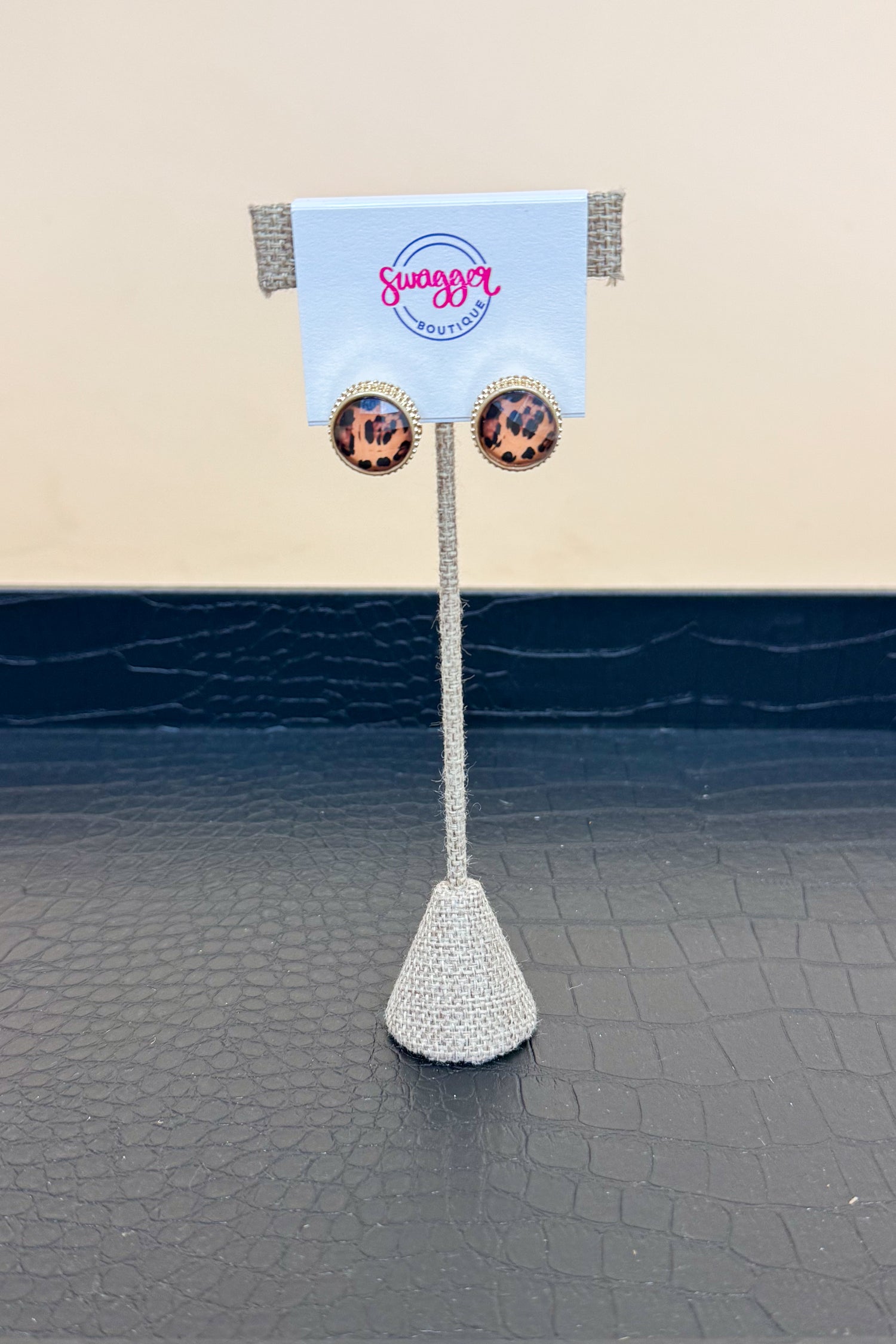 Pair of leopard earrings on a decorative stand with a branded card in the background.