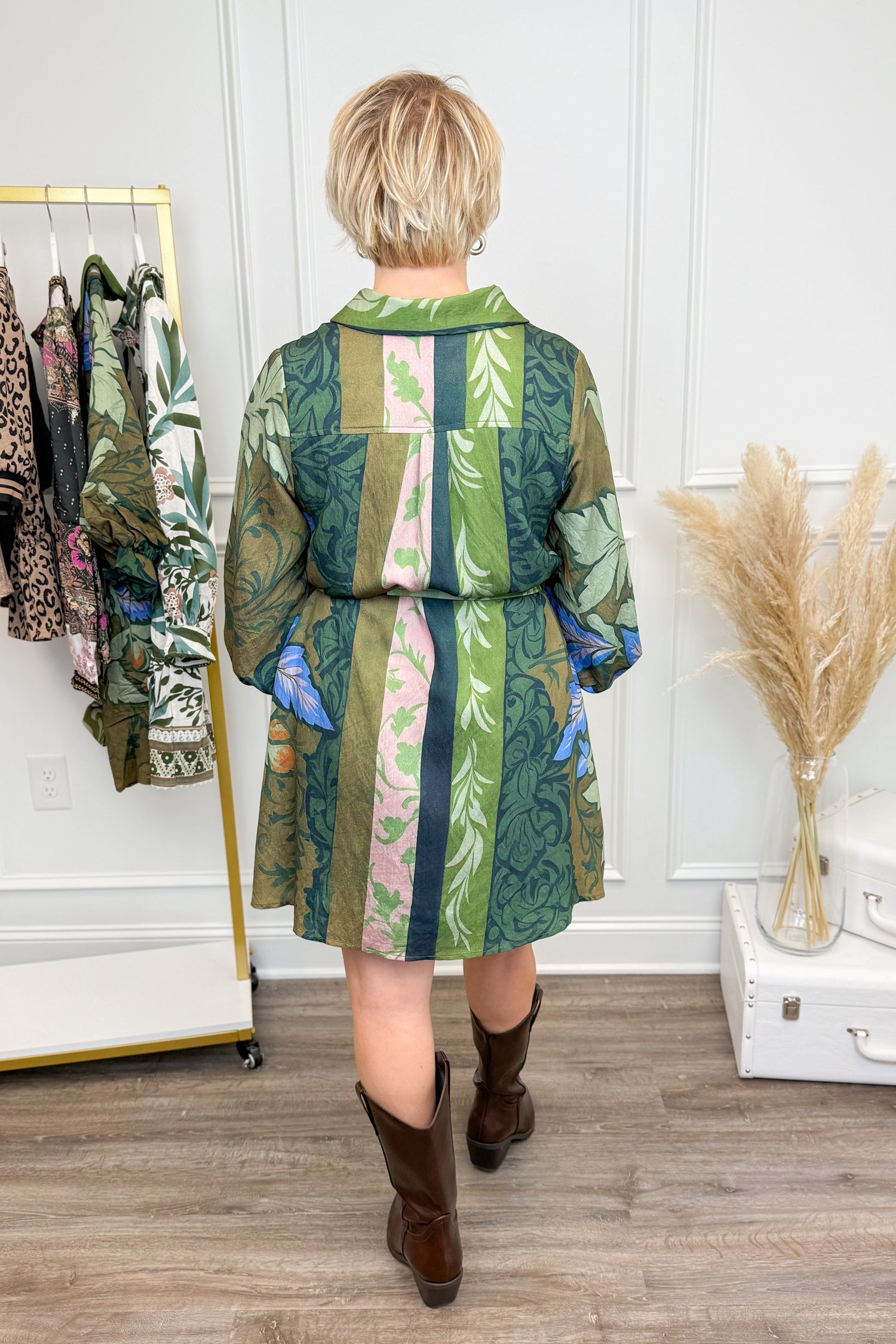 Person wearing a green and brown patterned dress, standing in a room with a rack of clothes and decorative items.