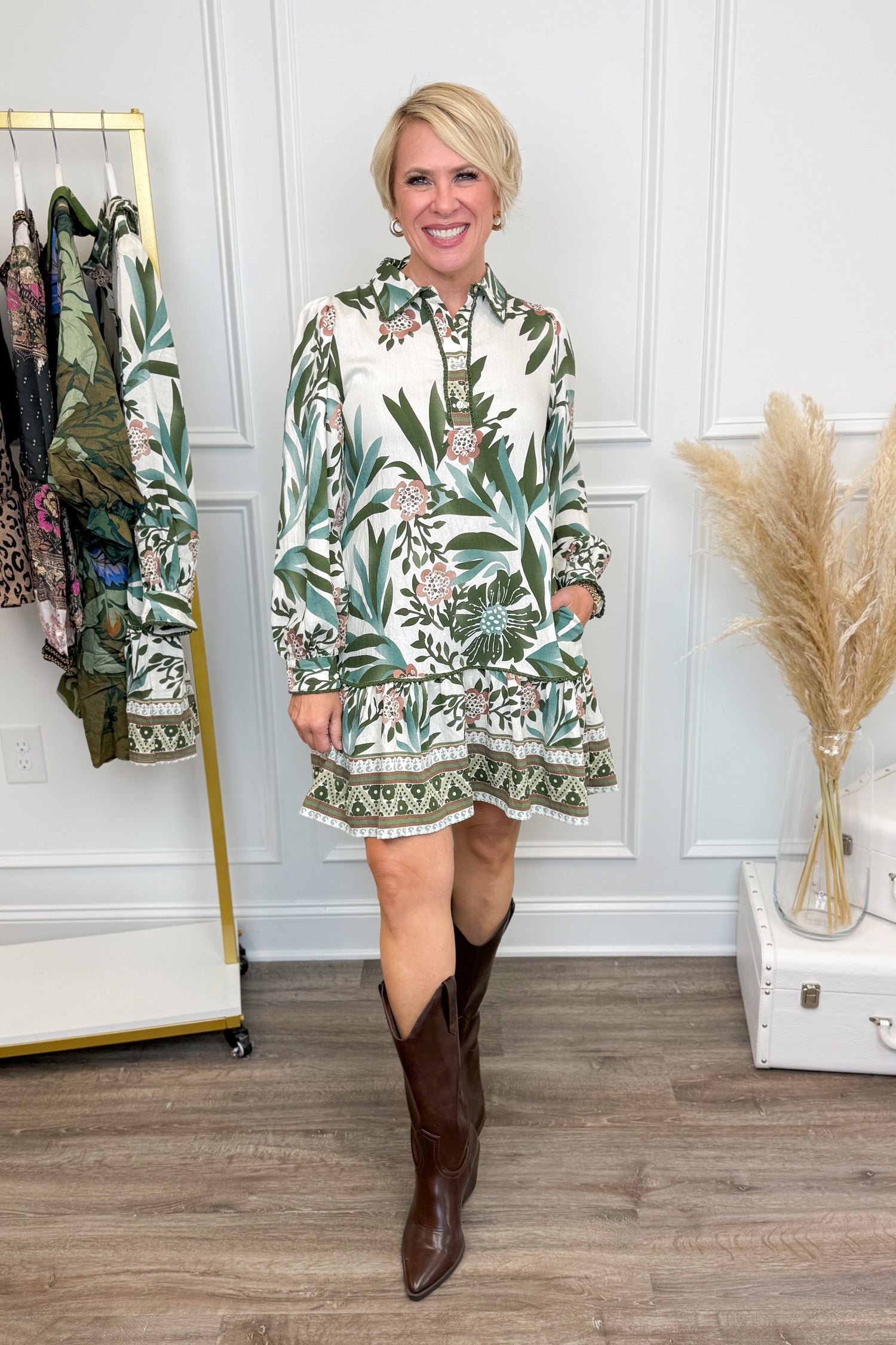 Woman wearing a floral dress and brown boots in a room with a white wall and wooden floor.