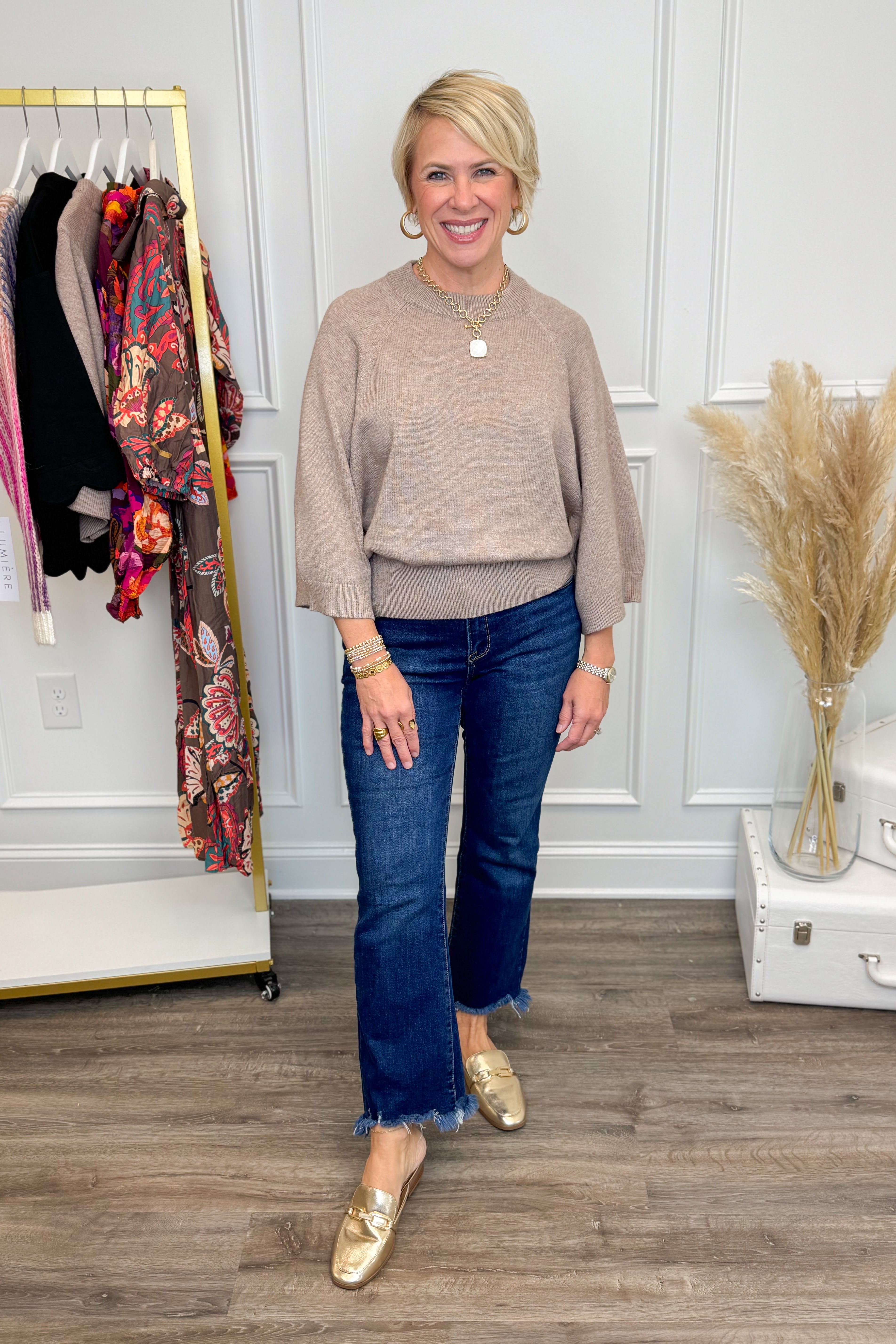 Woman wearing a beige sweater and blue jeans standing in a room with a rack of clothes and decorative elements.