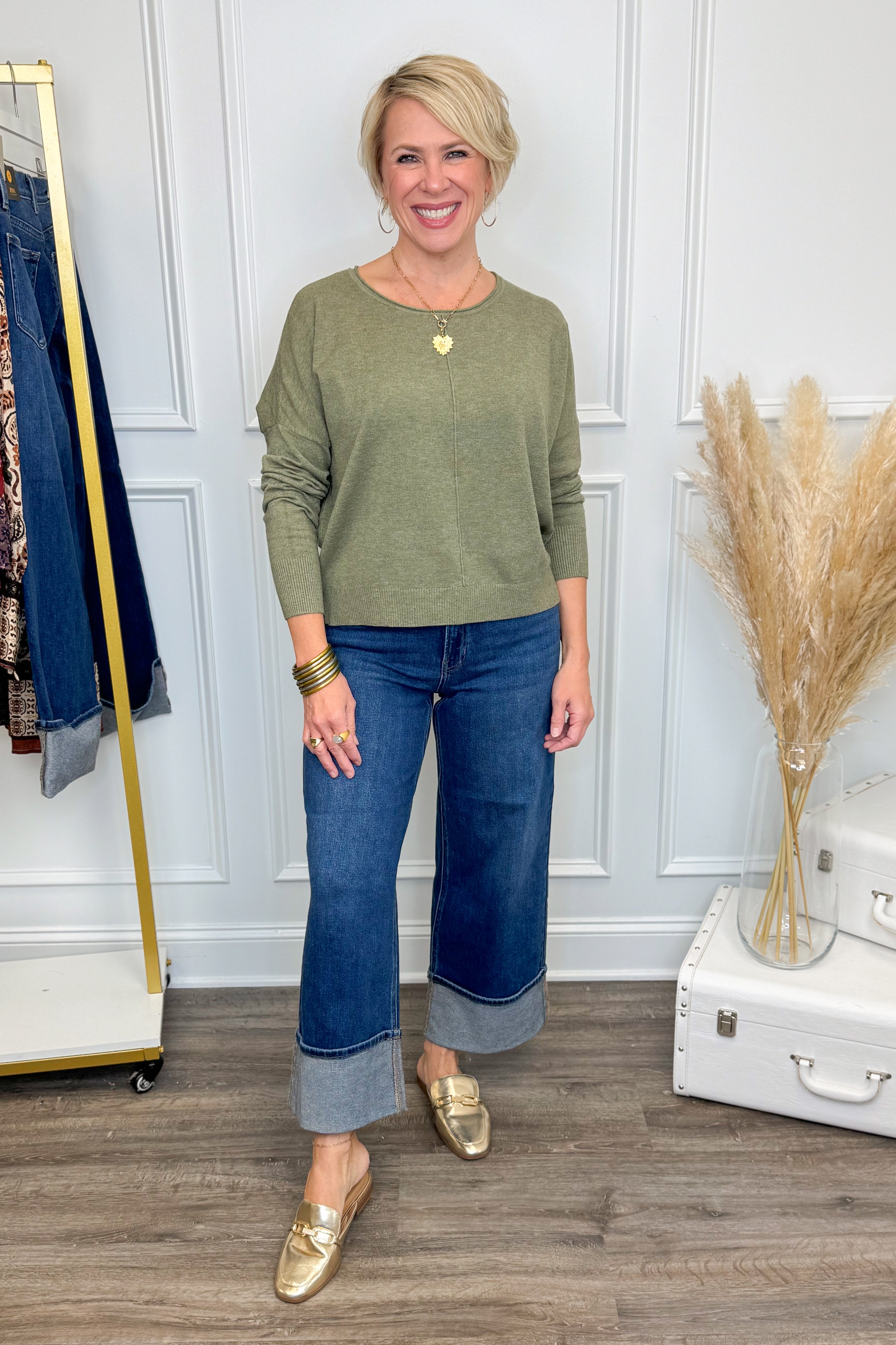 Woman wearing a green sweater and blue jeans standing in a room with a white wall and wooden floor.