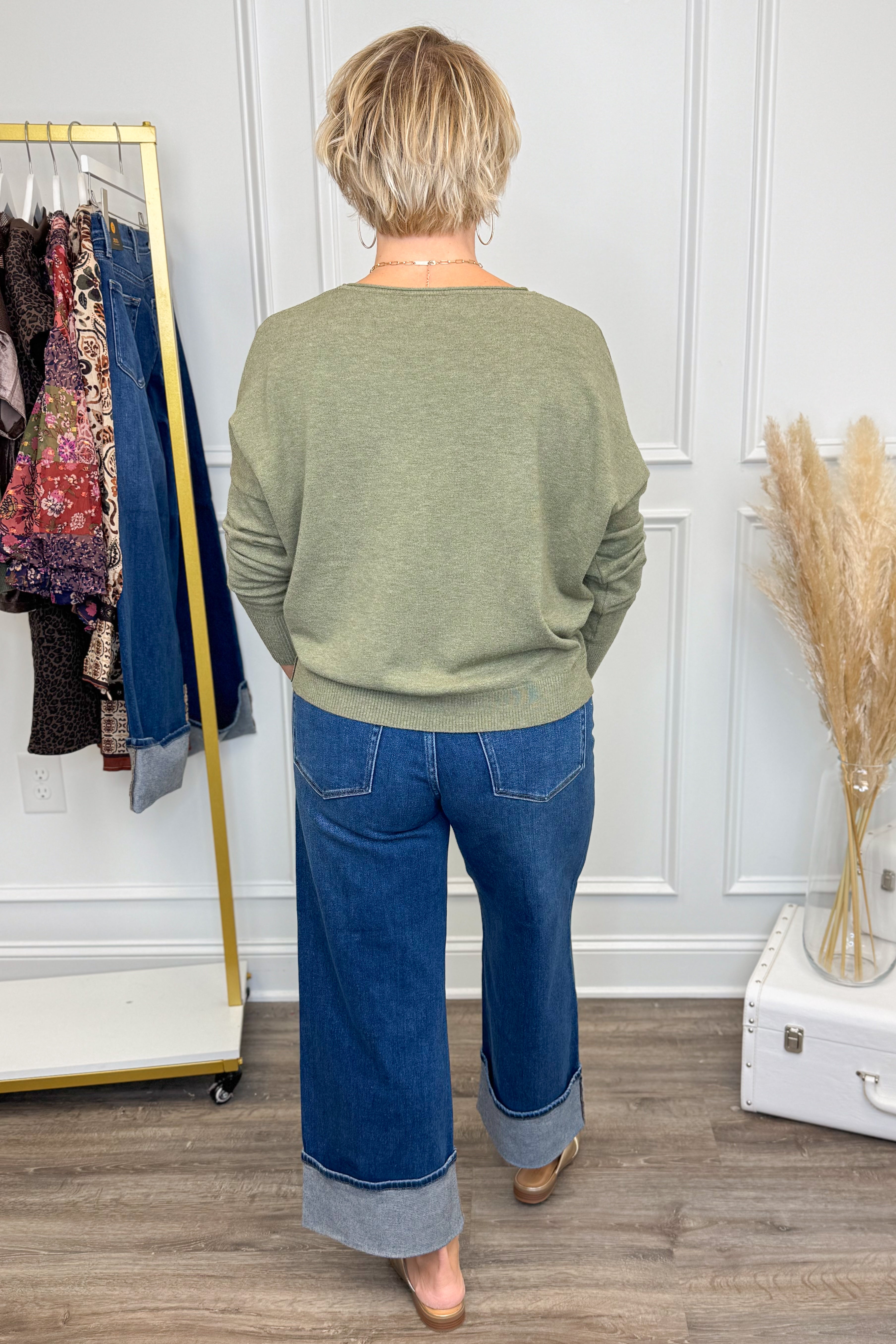 Person wearing a green sweater and blue jeans standing in a room with a rack of clothes and a suitcase.