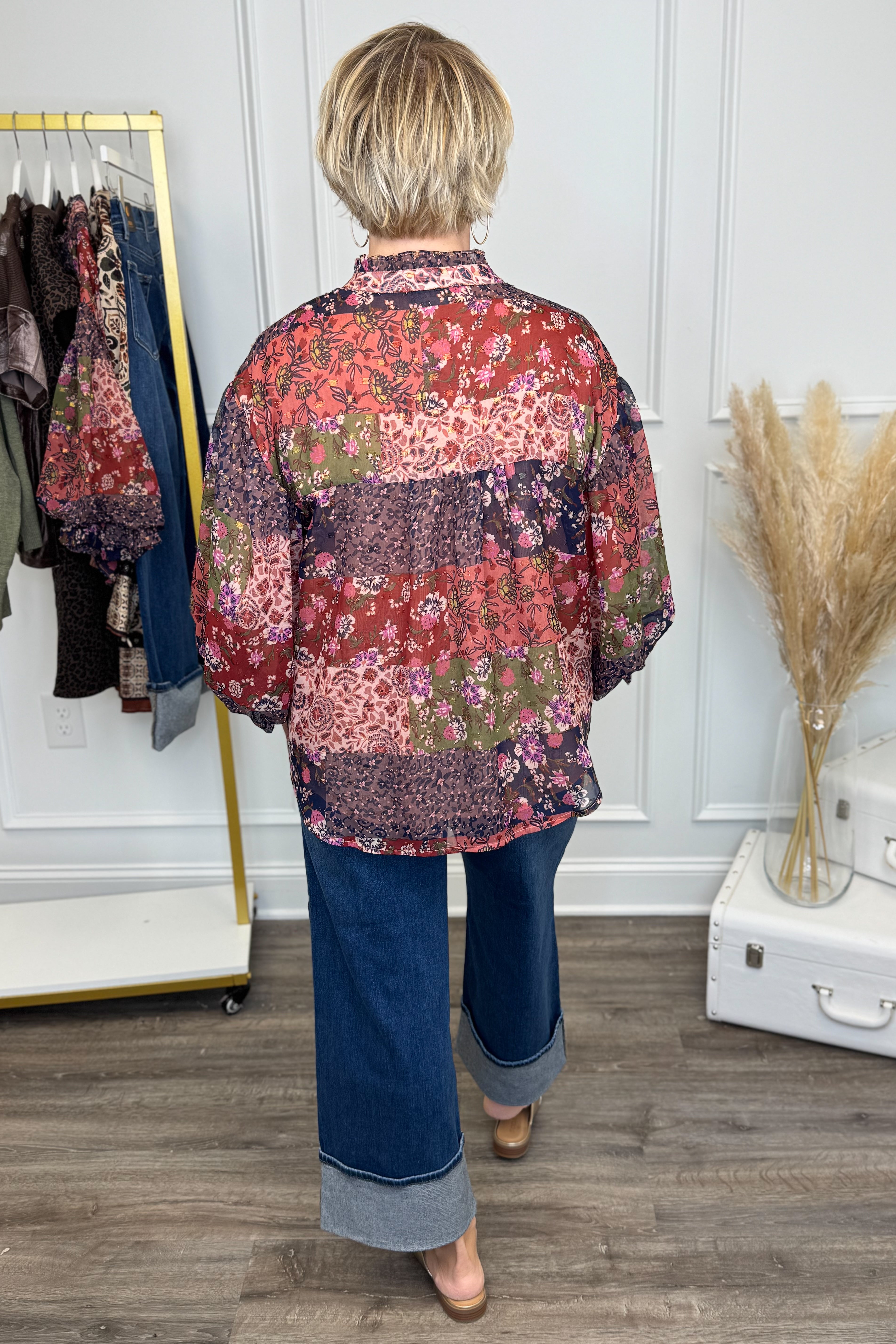 Person wearing a colorful patchwork shirt and blue jeans in a room with a clothing rack and decorative items.
