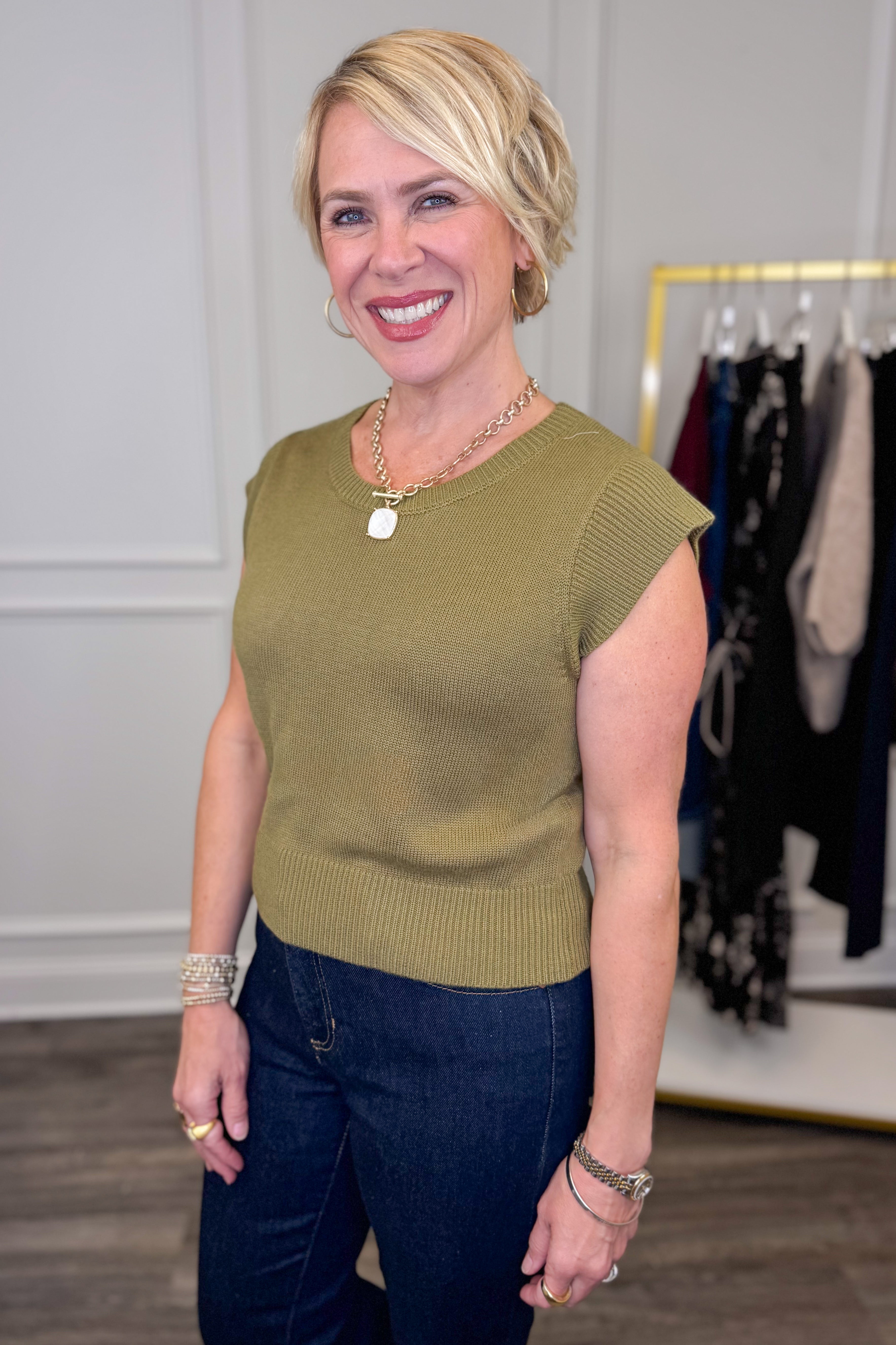 Woman wearing a green sleeveless sweater and dark pants in a clothing store.