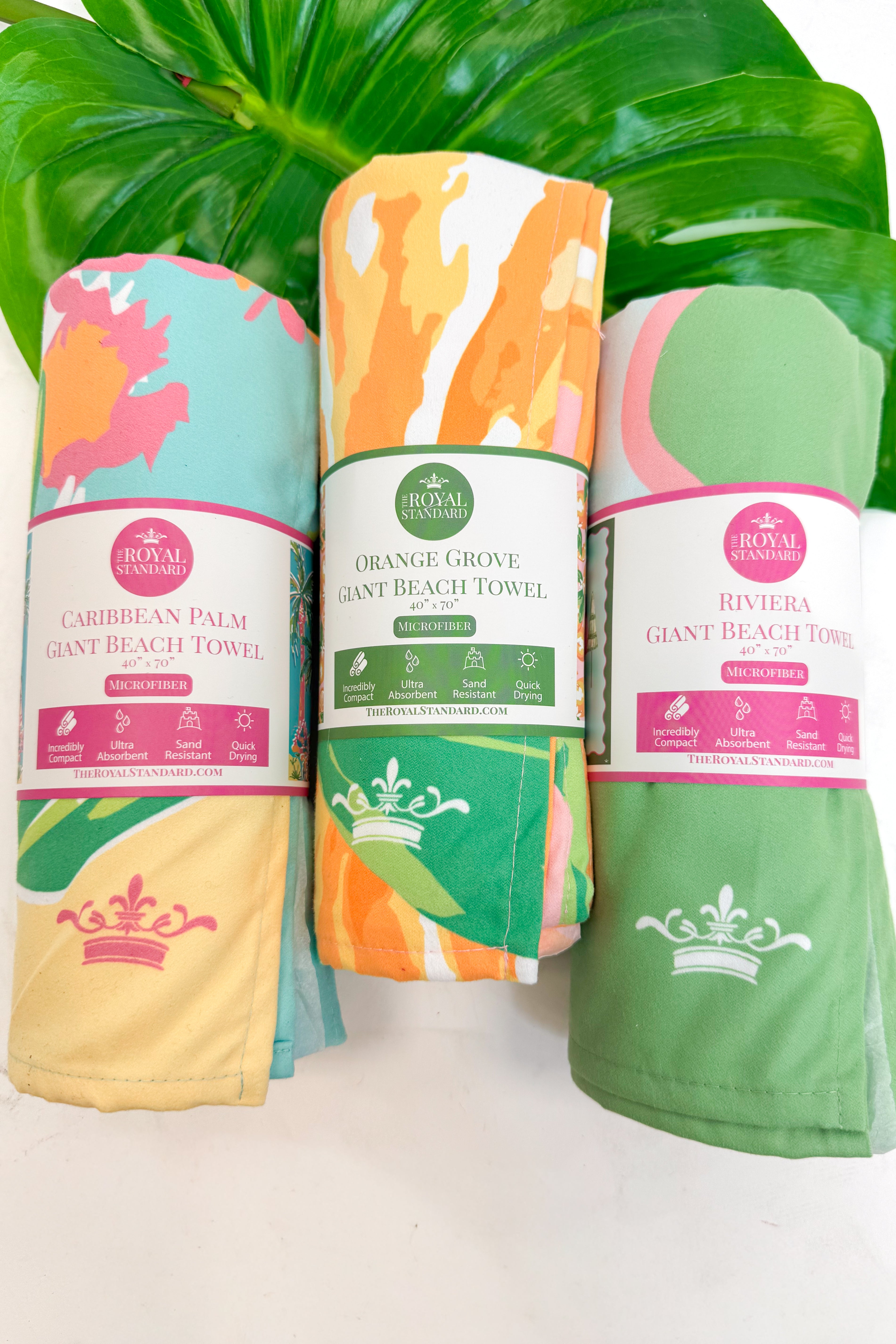 Three colorful beach towels with brand labels on a white surface with green leaves in the background.