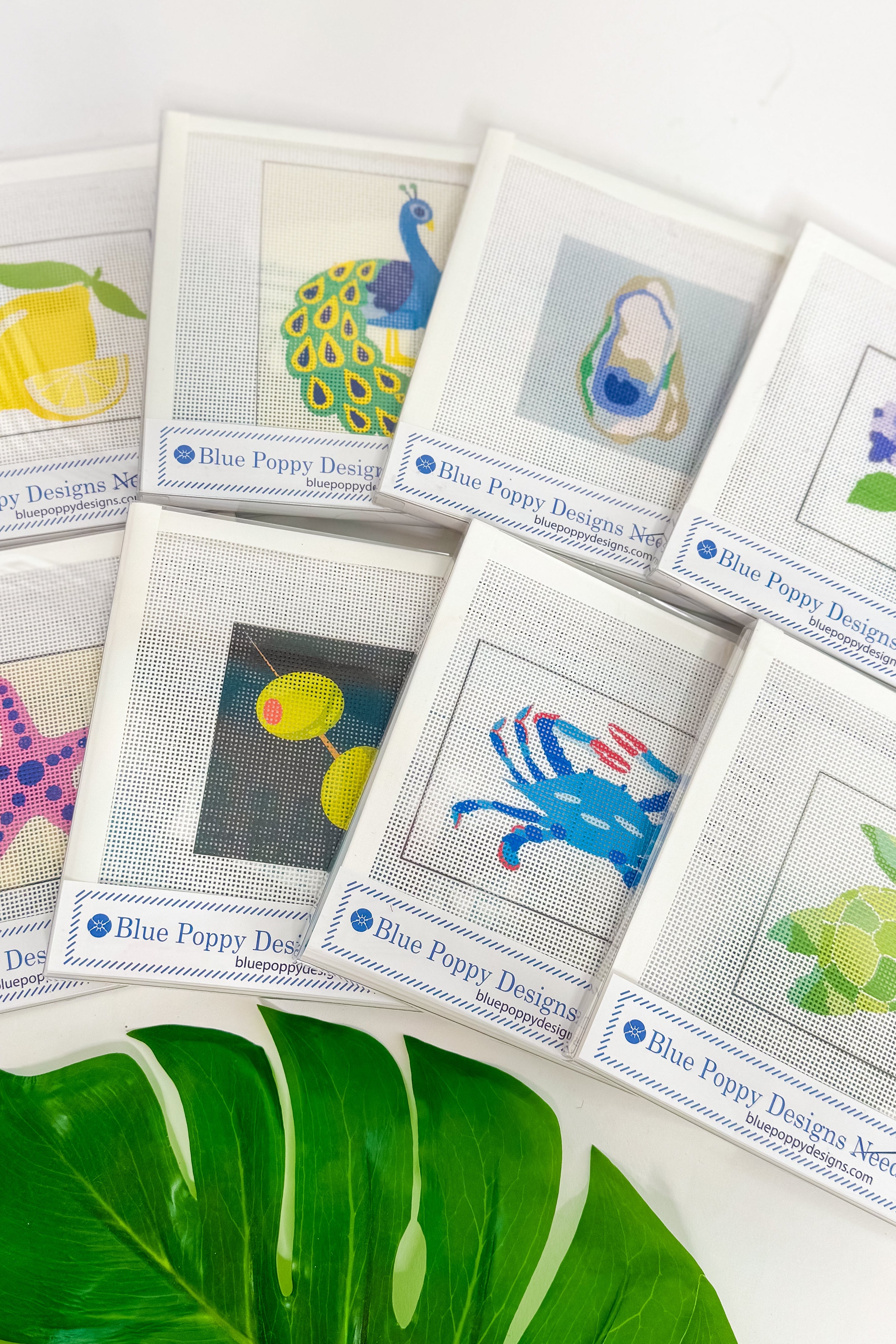 Collection of cross-stitch pattern cards by Blue Poppy Designs on a white surface with green leaves.