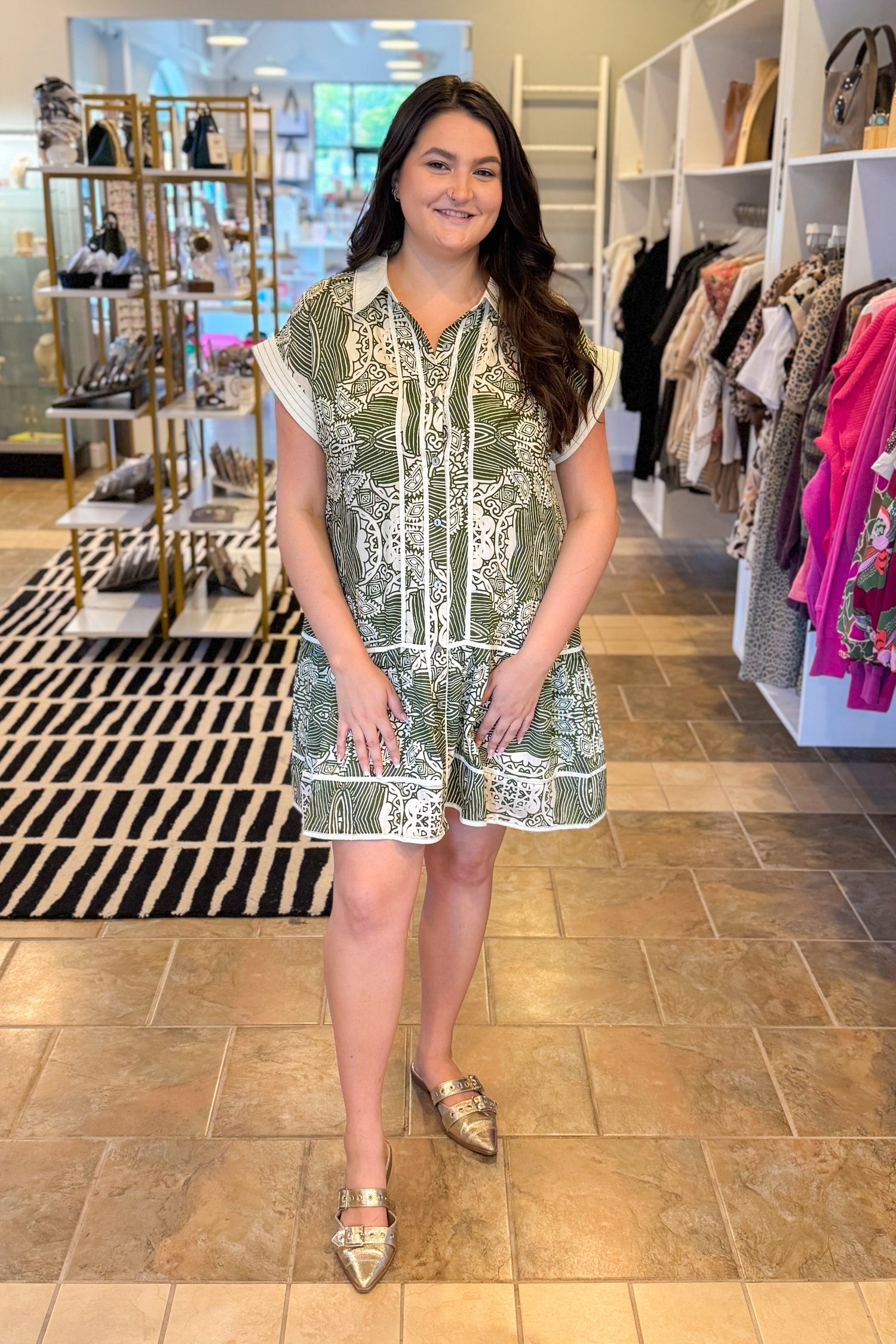 Woman in a patterned dress standing in a store with clothing racks and shoes in the background.