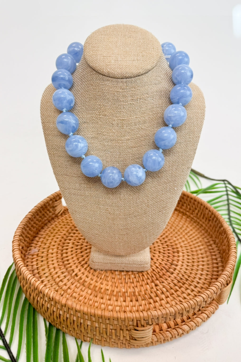 Blue beaded necklace on a beige mannequin bust with a woven basket and green leaves in the background