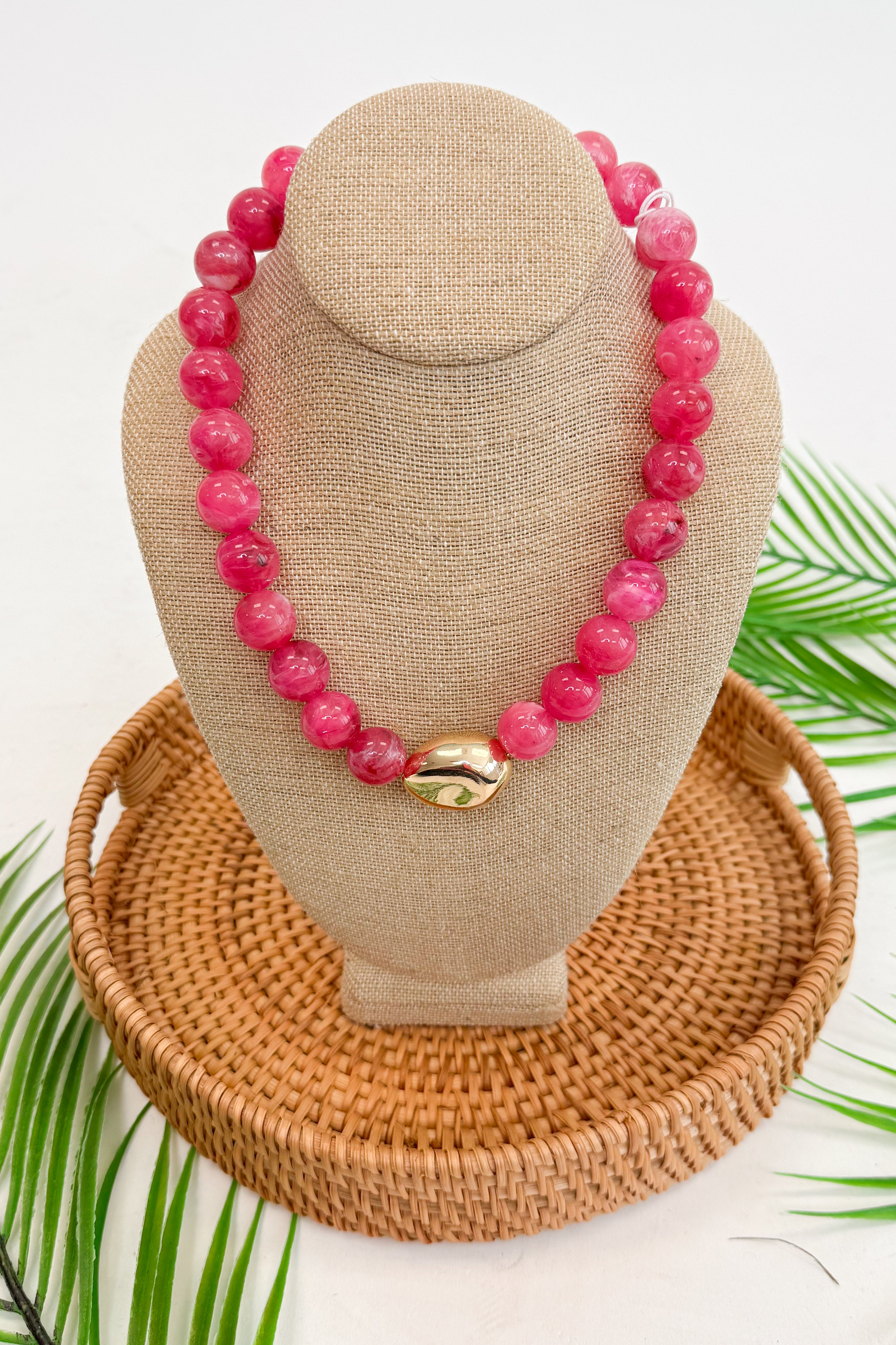 Pink beaded necklace on a beige bust with a woven base, surrounded by green leaves.