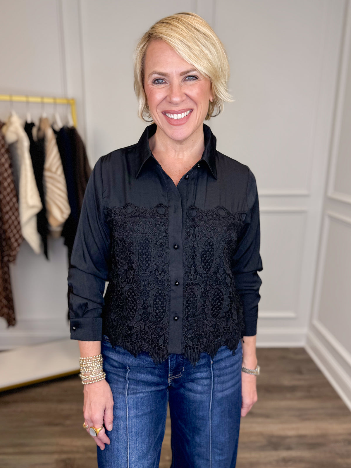 A smiling blonde woman wearing a black long-sleeve button-up blouse with a lace panel across the chest and blue jeans, standing in an indoor boutique setting.