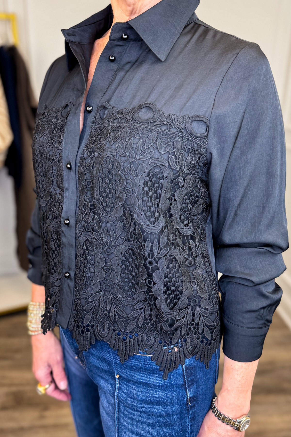 A person wearing a black long-sleeve button-up shirt with an intricate black lace overlay across the chest, paired with blue jeans.