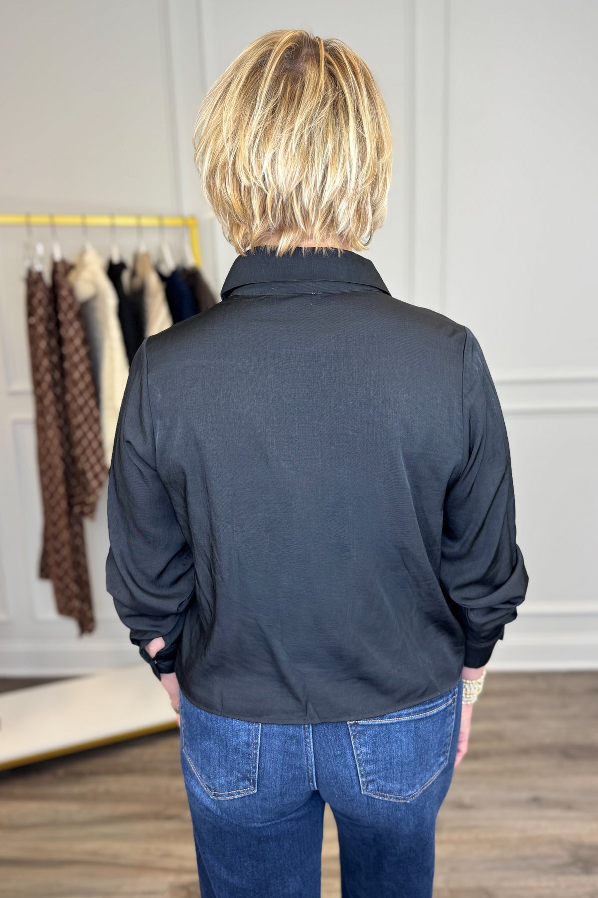 A woman viewed from behind wearing a black long-sleeve collared shirt and blue jeans in a boutique setting.