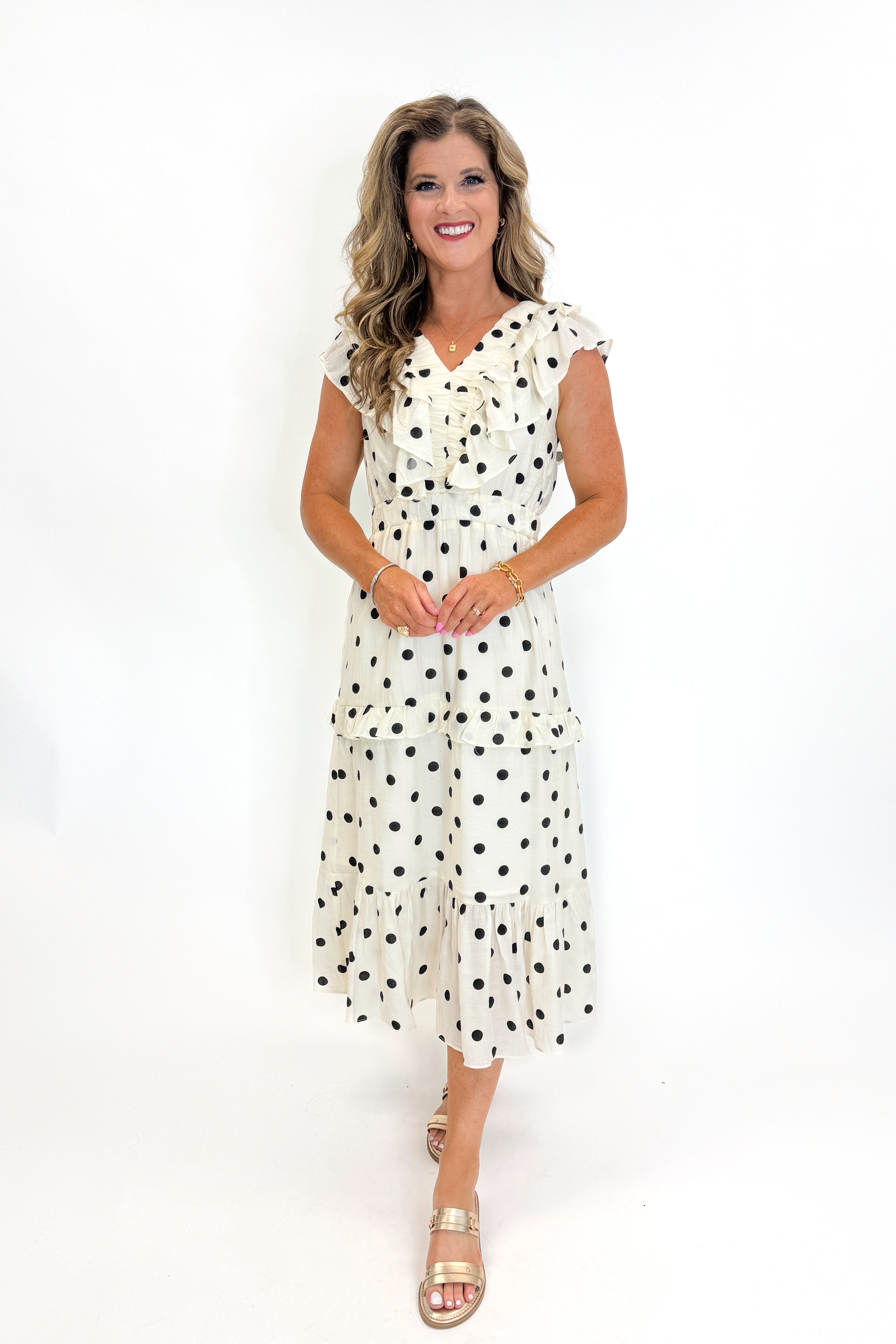 Front view of woman wearing Swagger Boutiqueβs Ivory & Black Polka Dot Ruffle Dress with sandals against white studio background.