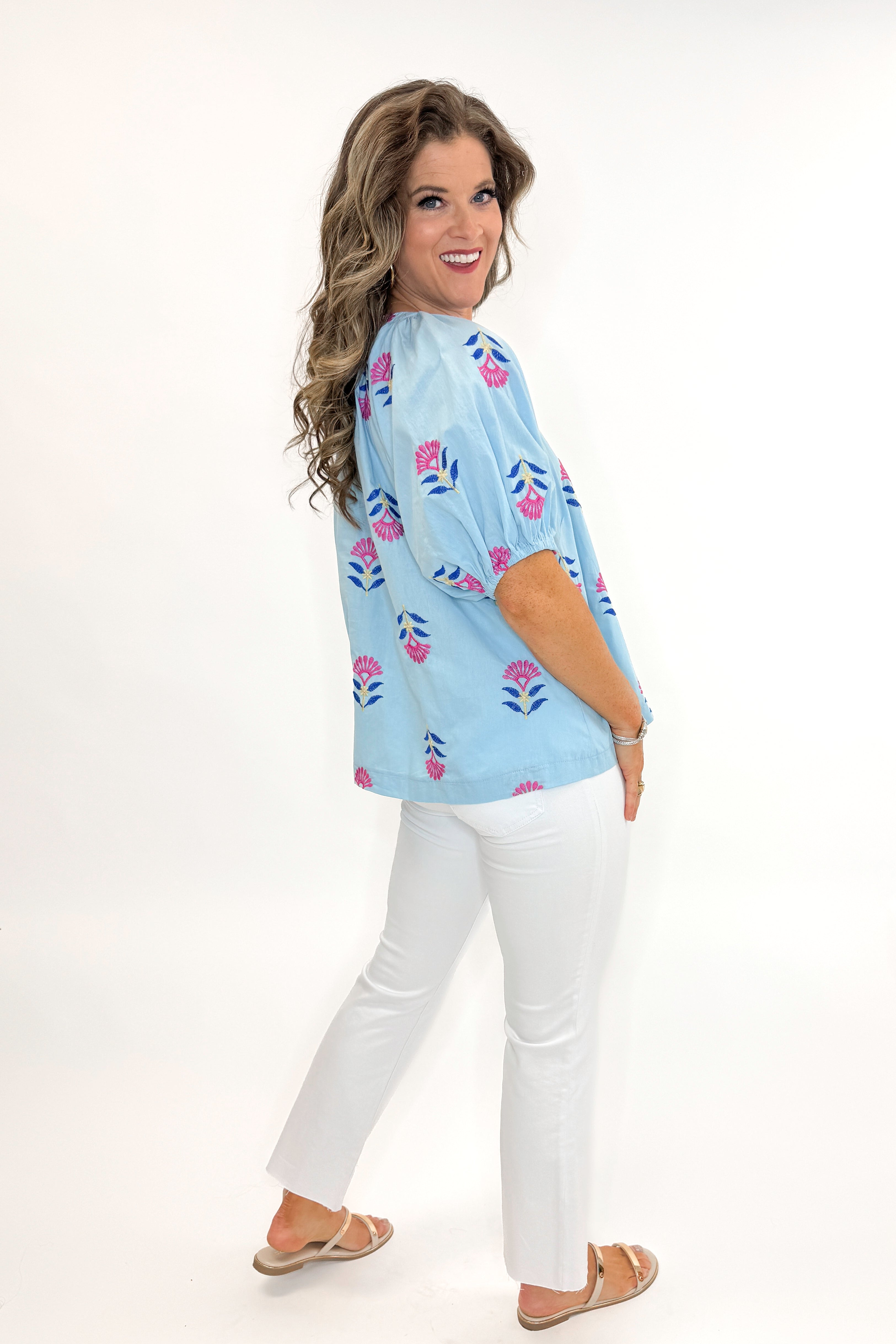 Side view of woman wearing Blue & Multi Floral Embroidered Top with jeans against white studio background.