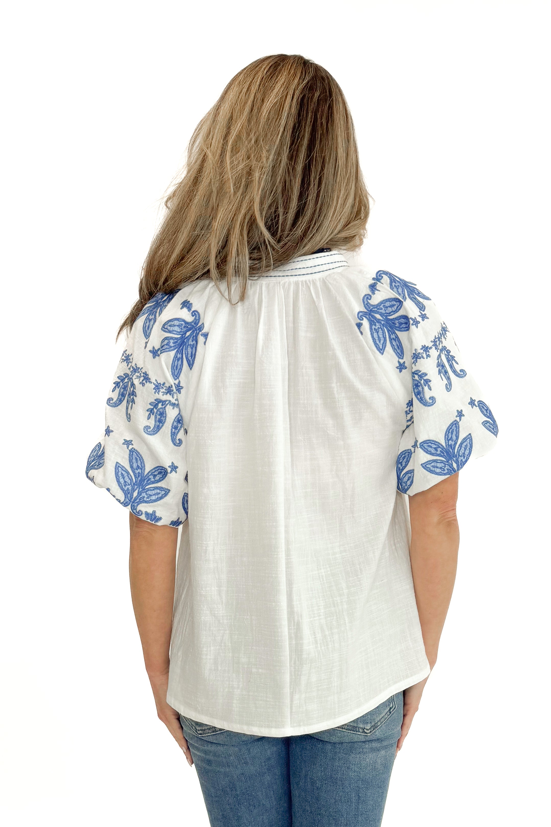 Back view of woman wearing Off White With Blue Embroidered Sleeve Top with jeans against white studio background.