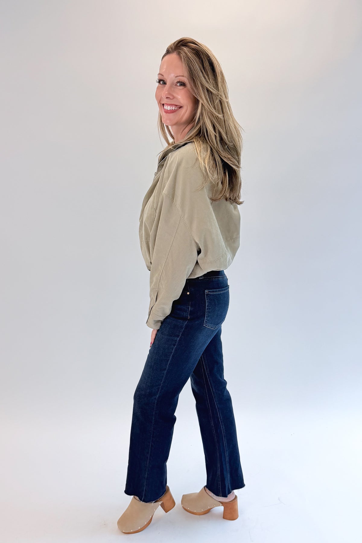Side view of woman wearing Swagger Boutique’s Khaki Bubble Hem Bomber Jacket with jeans against white studio background.