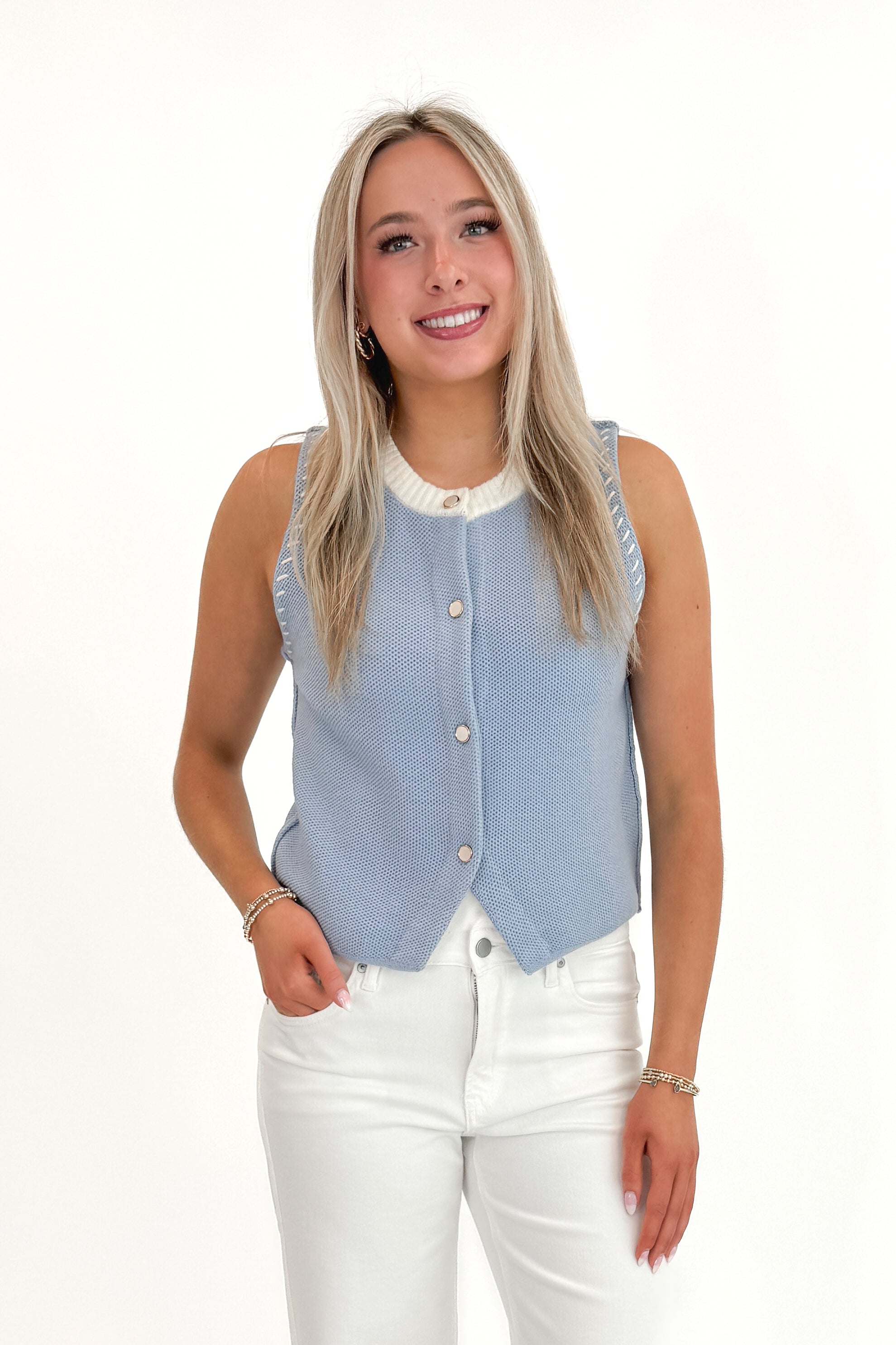 Front view of woman wearing Light Blue Button Up Sleeveless Sweater with white jeans against white studio background.