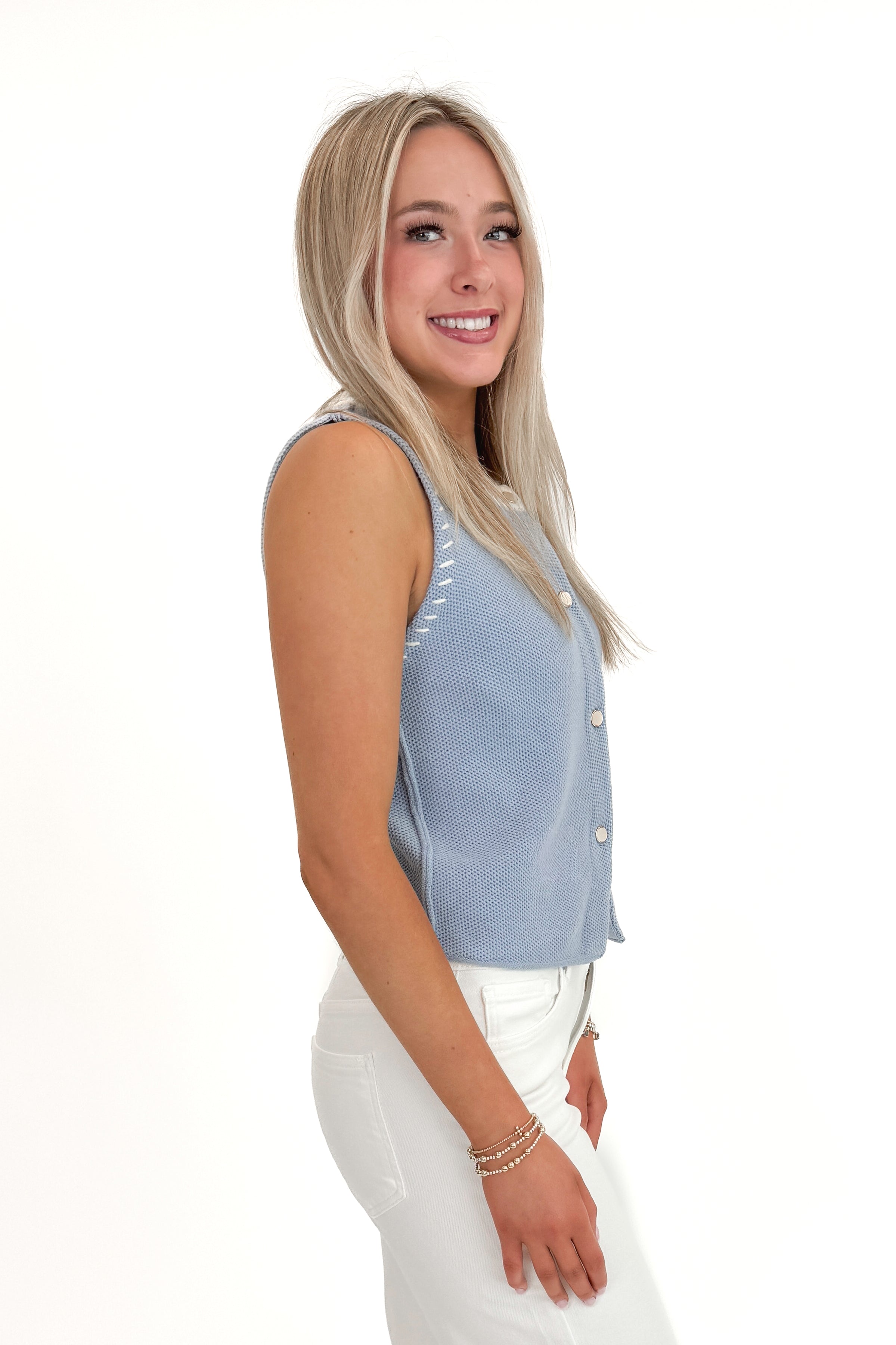 Side view of woman wearing Light Blue Button Up Sleeveless Sweater with white jeans against white studio background.