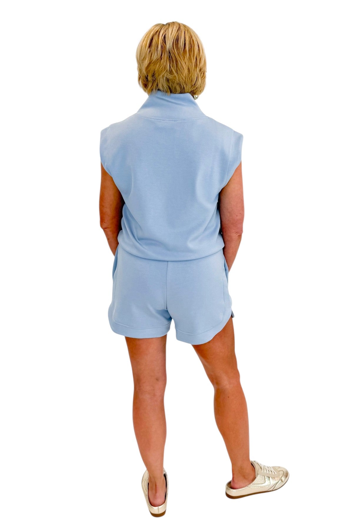 Back view of woman wearing Swagger Boutiqueās Light Blue Sleeveless 1/4 Zip Top with matching shorts against white studio background.