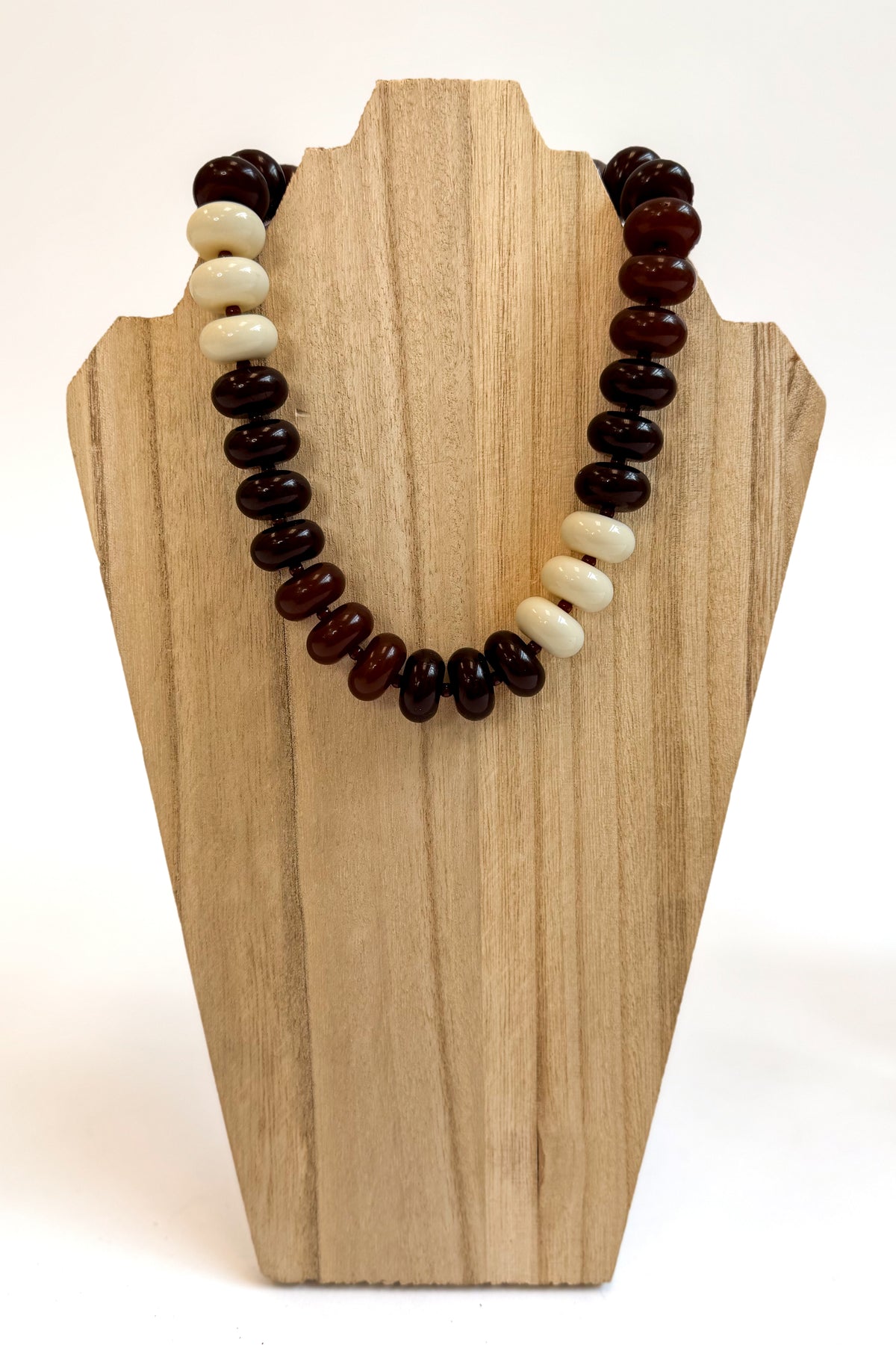 A beaded necklace with alternating dark brown and cream beads, displayed on a light wood stand against a white background.