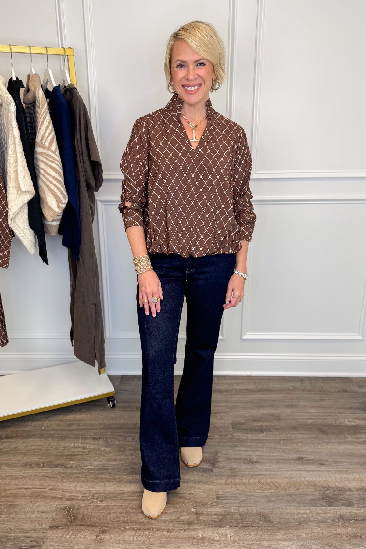 A woman modeling a brown and white diamond-patterned button-down blouse with dark blue flare jeans and beige heels, standing in a room with a clothing rack and white paneled walls.