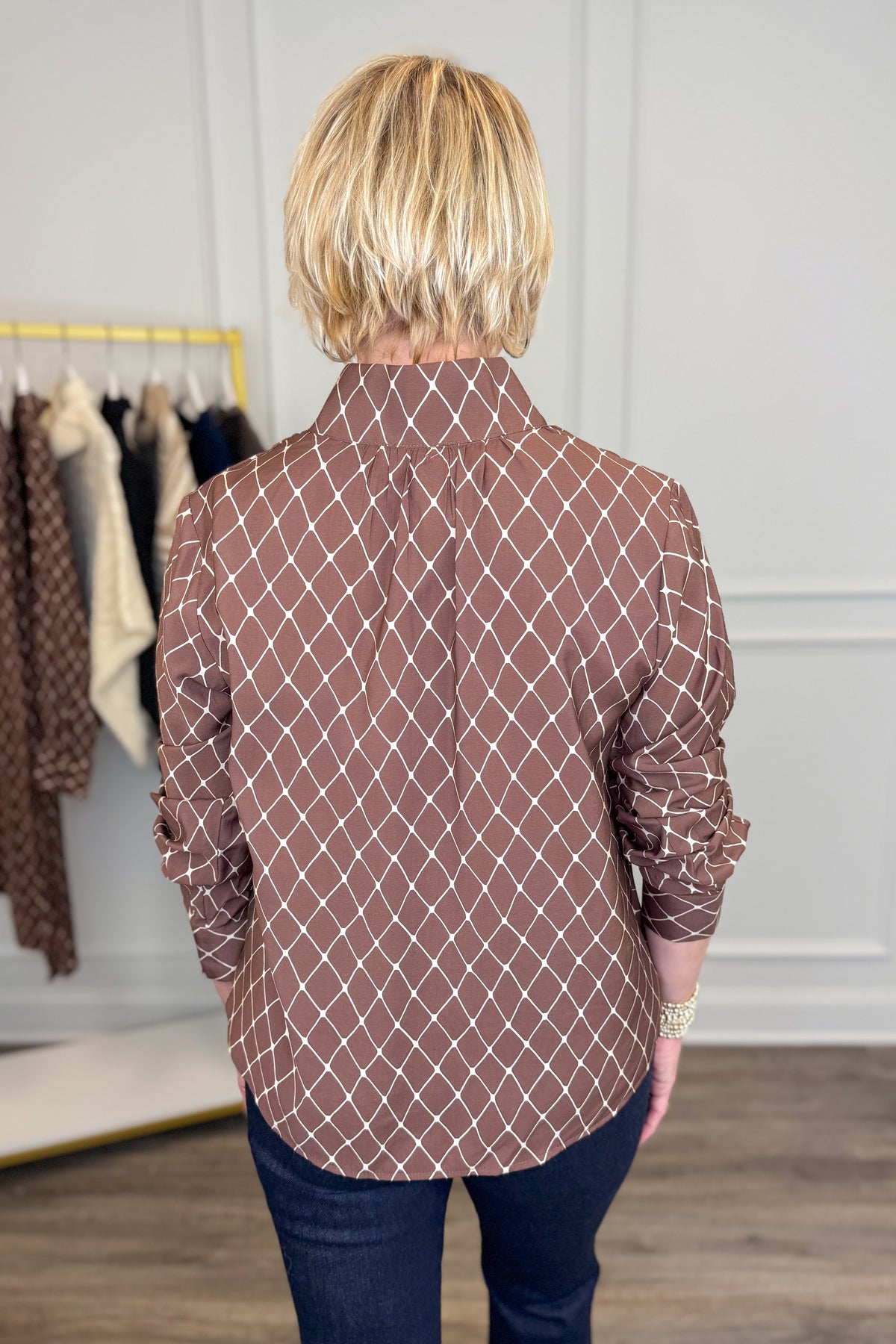 A woman viewed from behind wearing a brown long-sleeve button-up shirt with a white geometric diamond pattern, paired with dark blue jeans in an indoor boutique setting.