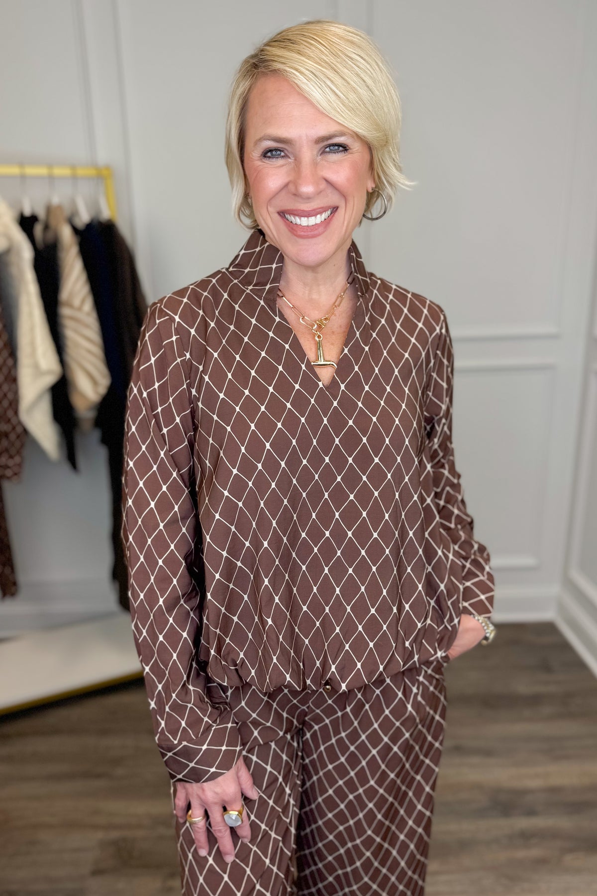 A smiling blonde woman wearing a brown and beige diamond-patterned long-sleeve top and matching pants, standing in a boutique setting with a clothing rack visible in the background.